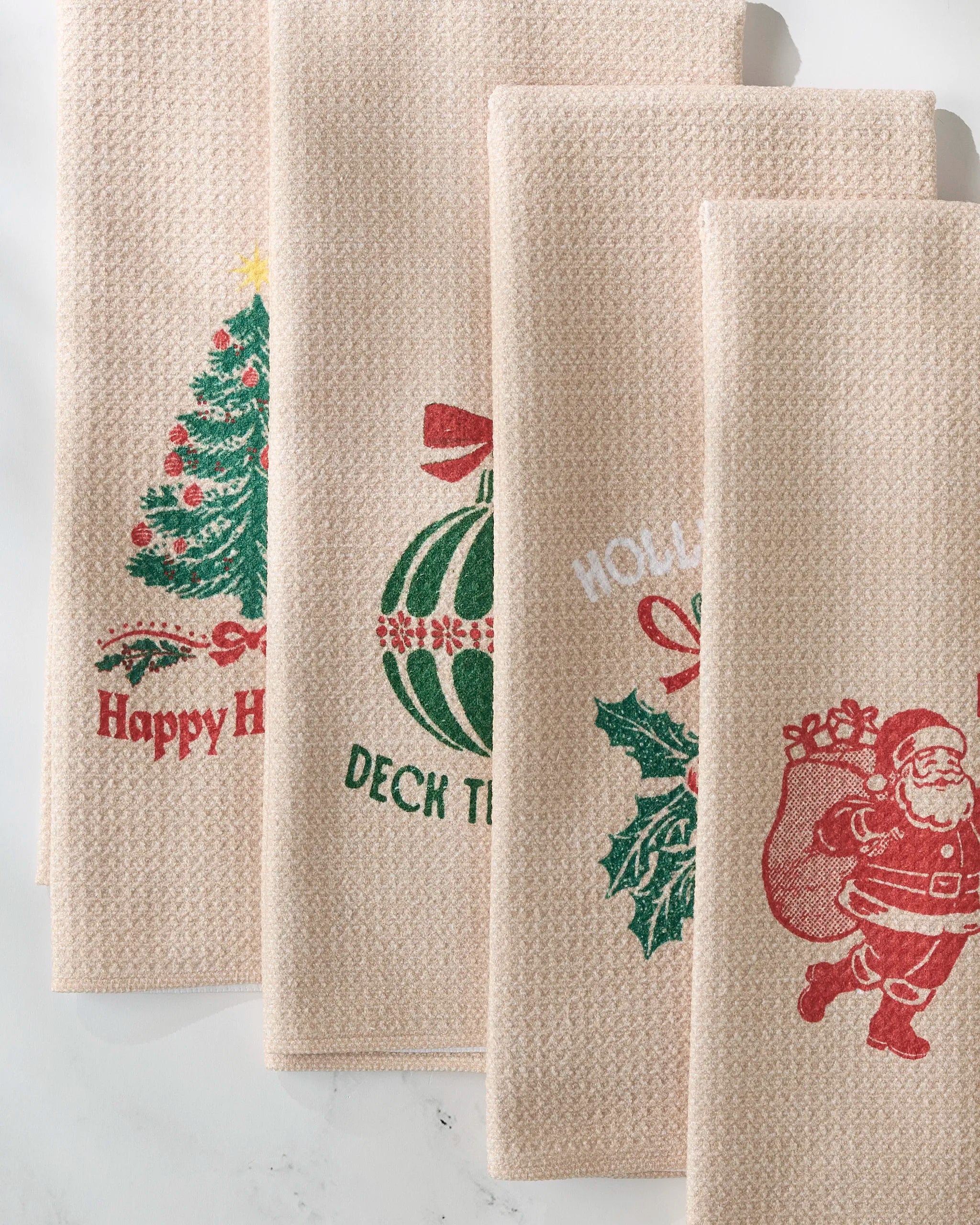 The GEOMETRY Festive Charm set includes four beige kitchen towels, each featuring a unique holiday design: Christmas tree, green ornament, holly leaves, and Santa Claus with a gift sack.