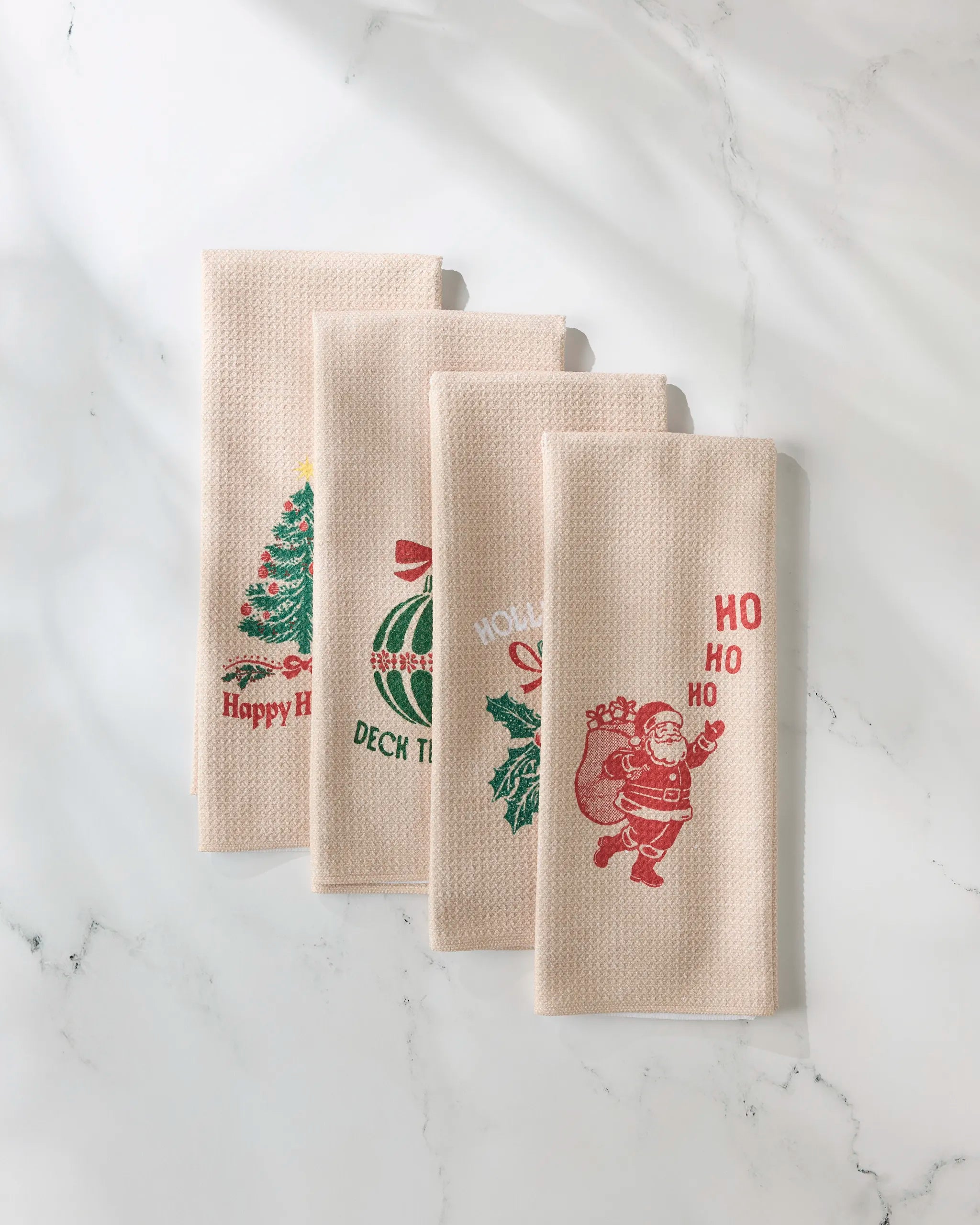 Four GEOMETRY Festive Charm beige kitchen towels are shown side by side on marble, each featuring a unique Christmas design: tree, ornaments, wreath, and Santa Claus with "HO HO HO.