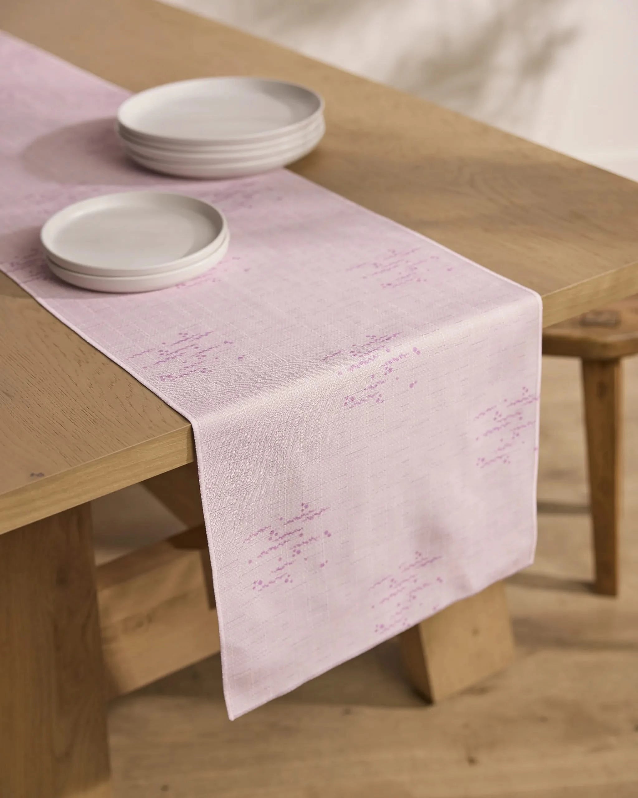 The Geometry Bubble Ditzy Dusty Rose stain-resistant table runner, featuring subtle purple stitching, lies on a wooden dining table topped with a stack of white ceramic plates.