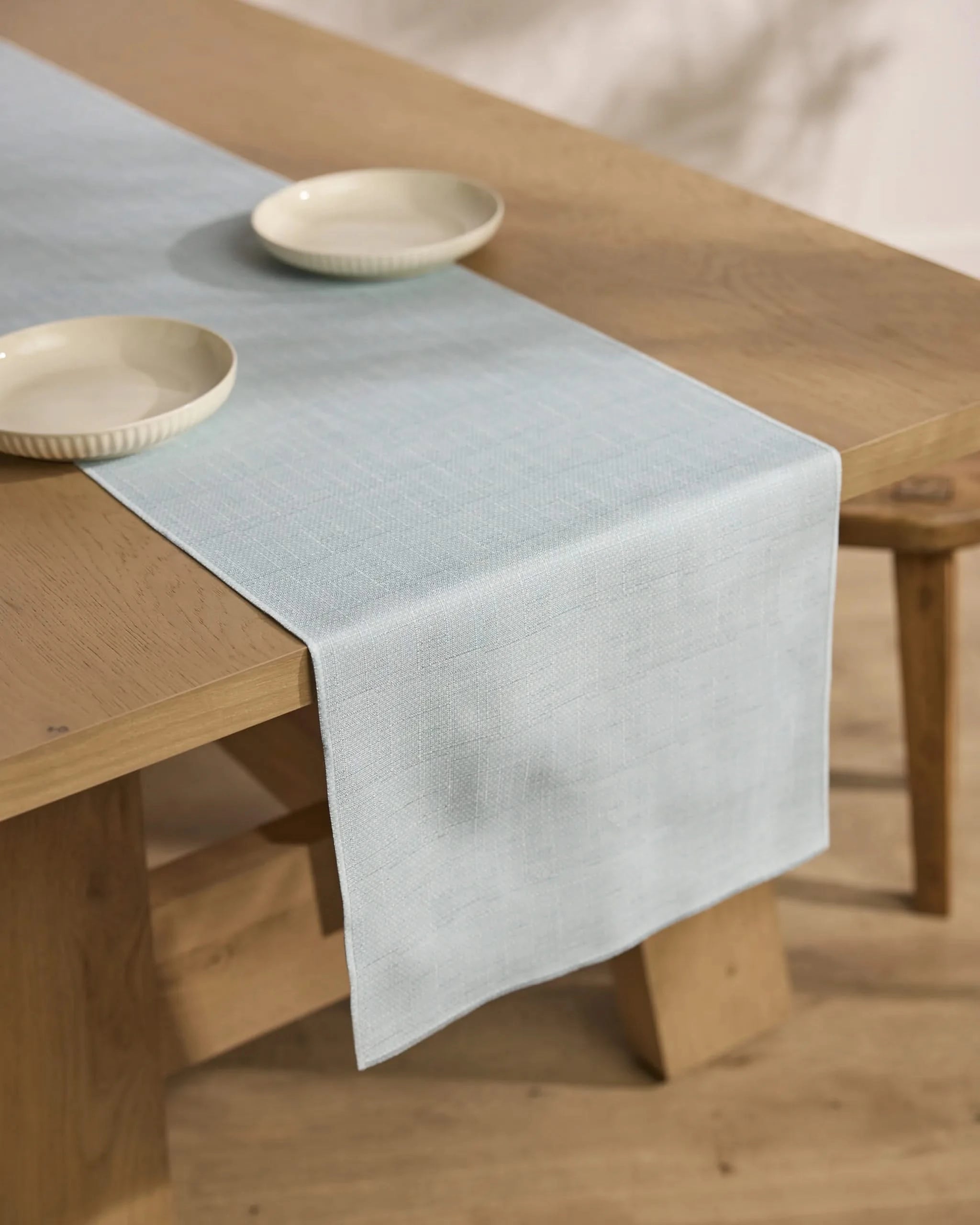 A Chambray Light Blue table runner by Geometry drapes over a wooden dining table topped with two cream-colored bowls. The setting is minimal, complemented by light wood flooring.