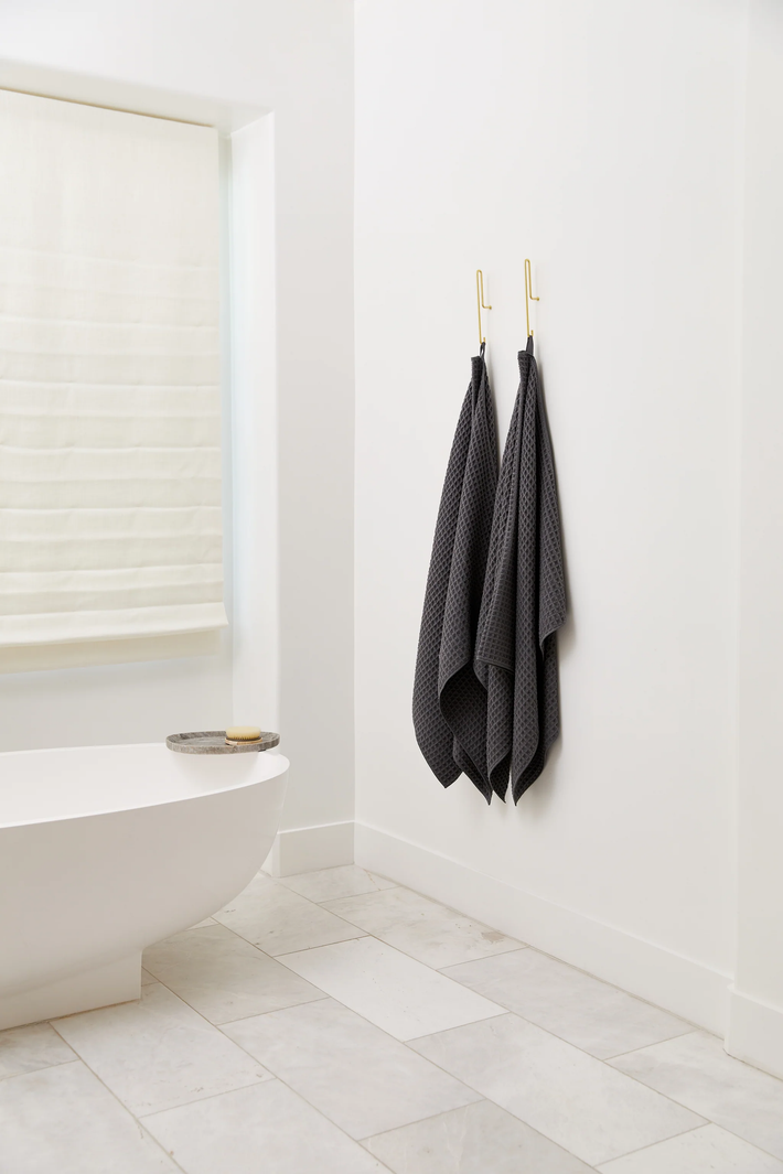A minimal, modern bathroom with white walls and tiled floor, featuring a white bathtub and window—plus the GEOMETRY Test Waffle Bundle in dark gray, your new favorite towels, hanging on gold hooks for a cozy, stylish touch.