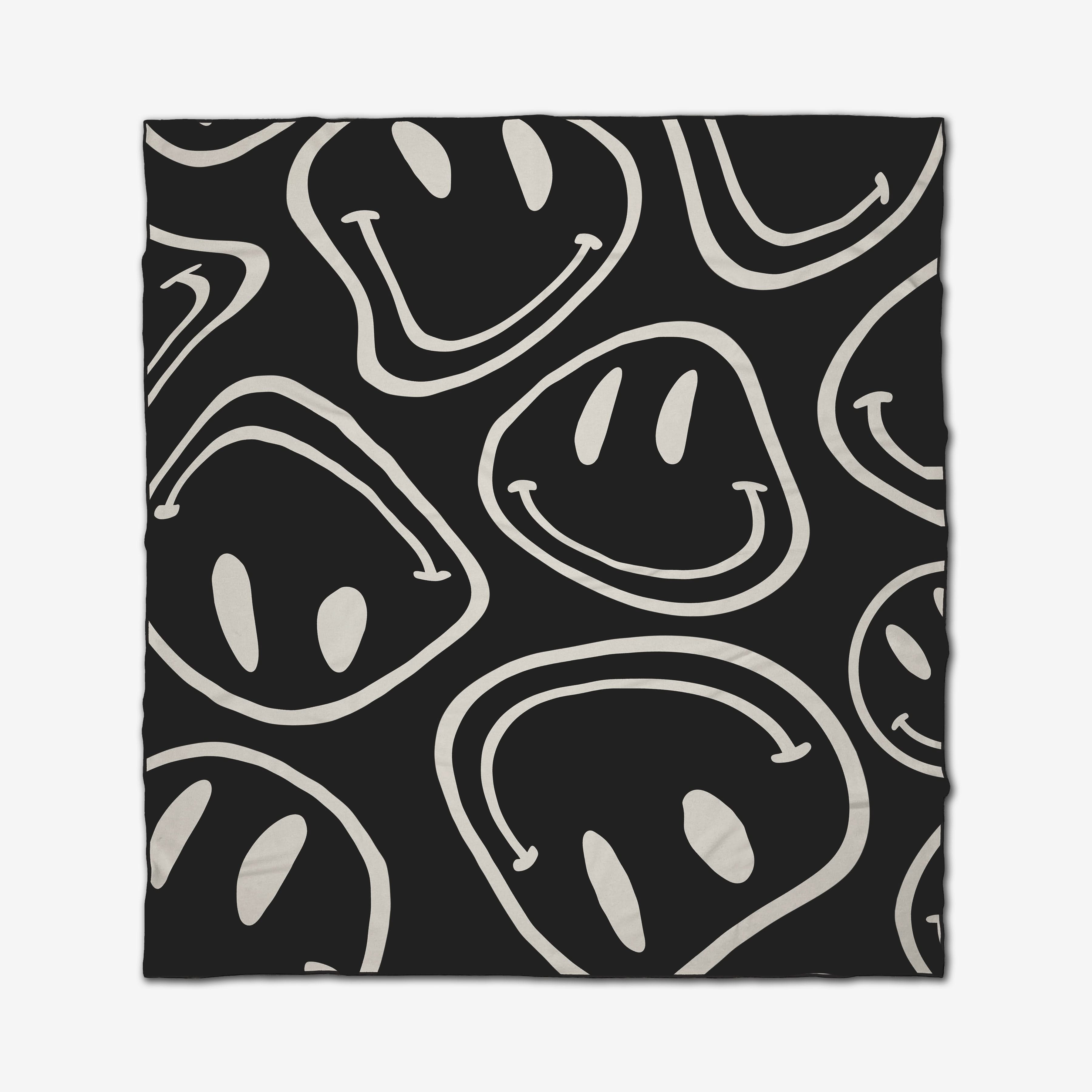 The GEOMETRY Mood Swing Dark beach blanket features large, white, playful hand-drawn smiley faces on a black background. Made from quick-drying recycled materials for eco-friendly fun in the sun.