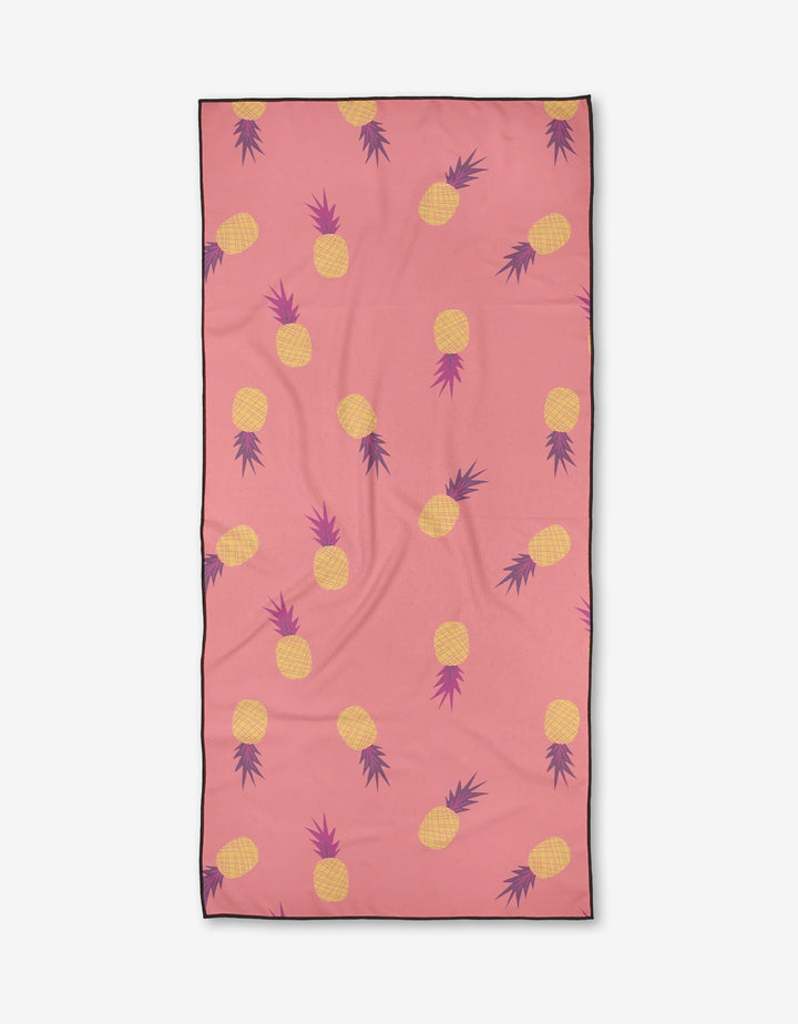 The Ananas by Jessica Swift is a compact beach towel featuring a pink background with repeating yellow pineapples, purple leaves, and a black border. Made from quick-drying fabric for easy use.