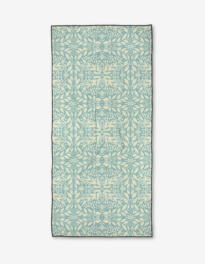 Bee Magic by Katie OShea is a rectangular micro-loop beach towel made from recycled materials, showcasing a pale blue and cream symmetrical leaf and vine pattern in an intricate botanical design on a white background.