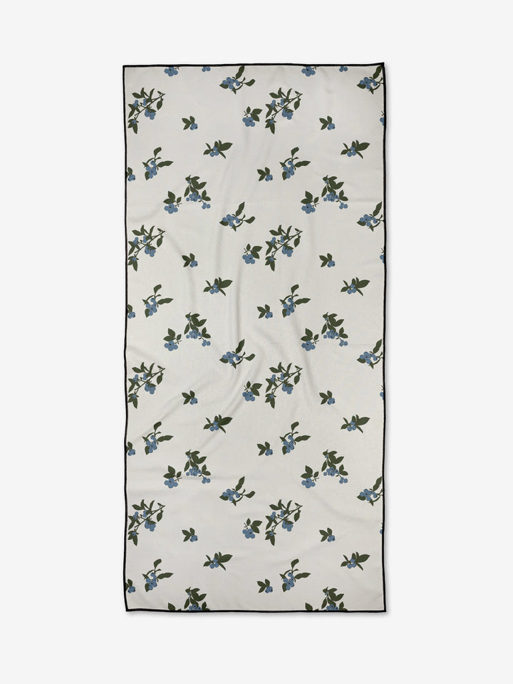 Aubrey Fairchild’s Blueberries is a rectangular white compact beach towel, adorned with scattered green leaves and small blue flowers, finished with a thin black border.
