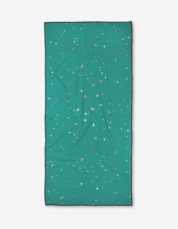 The Bailey Peyton Bubbly beach towel is rectangular, teal, and features a black border with colorful pink, yellow, white, and orange specks. Made from recycled materials and shown flat on a light background.