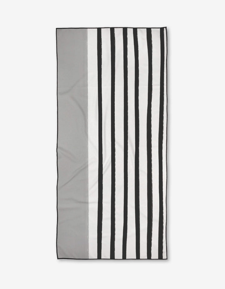 The Dawn beach towel by Kailee Wright is rectangular with a minimalist design—vertical black and white stripes on the right, solid gray on the left. Made from quick-drying recycled materials, its pictured against a plain white background.