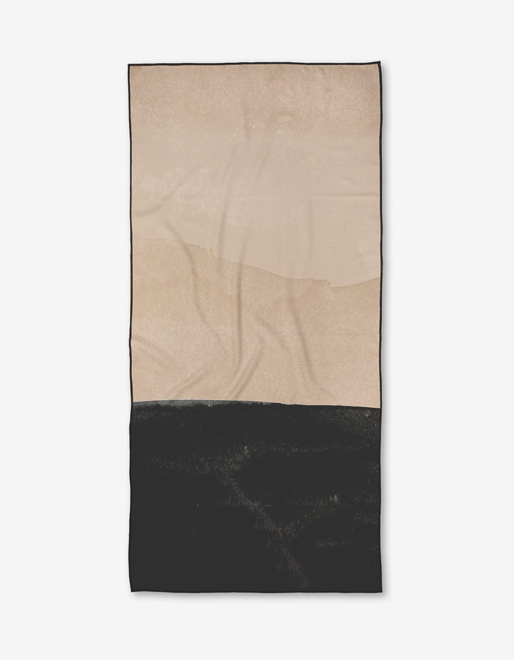 The Jojo towel by Kailee Wright is a rectangular, quick-drying towel made from recycled materials. It has a minimalist look with a beige upper half, solid black lower half separated by an uneven line, and naturally shows slight wrinkles.