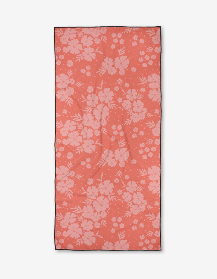 The Open Fields beach towel by Charlie Rowan Designs is rectangular, coral-colored, quick-drying, and features a light pink floral pattern of assorted flowers and leaves.