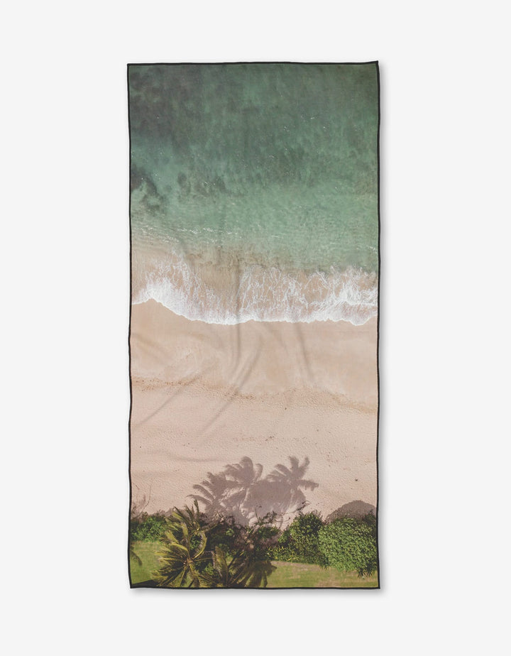 Paradise Found by Kait Hanson is a rectangular towel with an aerial beach print—turquoise waves, sandy shore, and palm shadows—crafted from ultra-absorbent, quick-drying recycled materials.