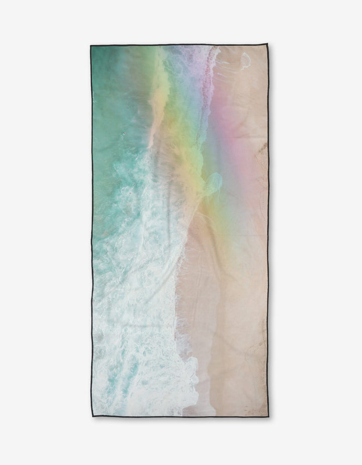The Rainbow Wave beach towel by Kait Hanson features an aerial wave design with a pastel rainbow across sand and water. Made from quick-drying recycled materials, it offers eco-friendly comfort.