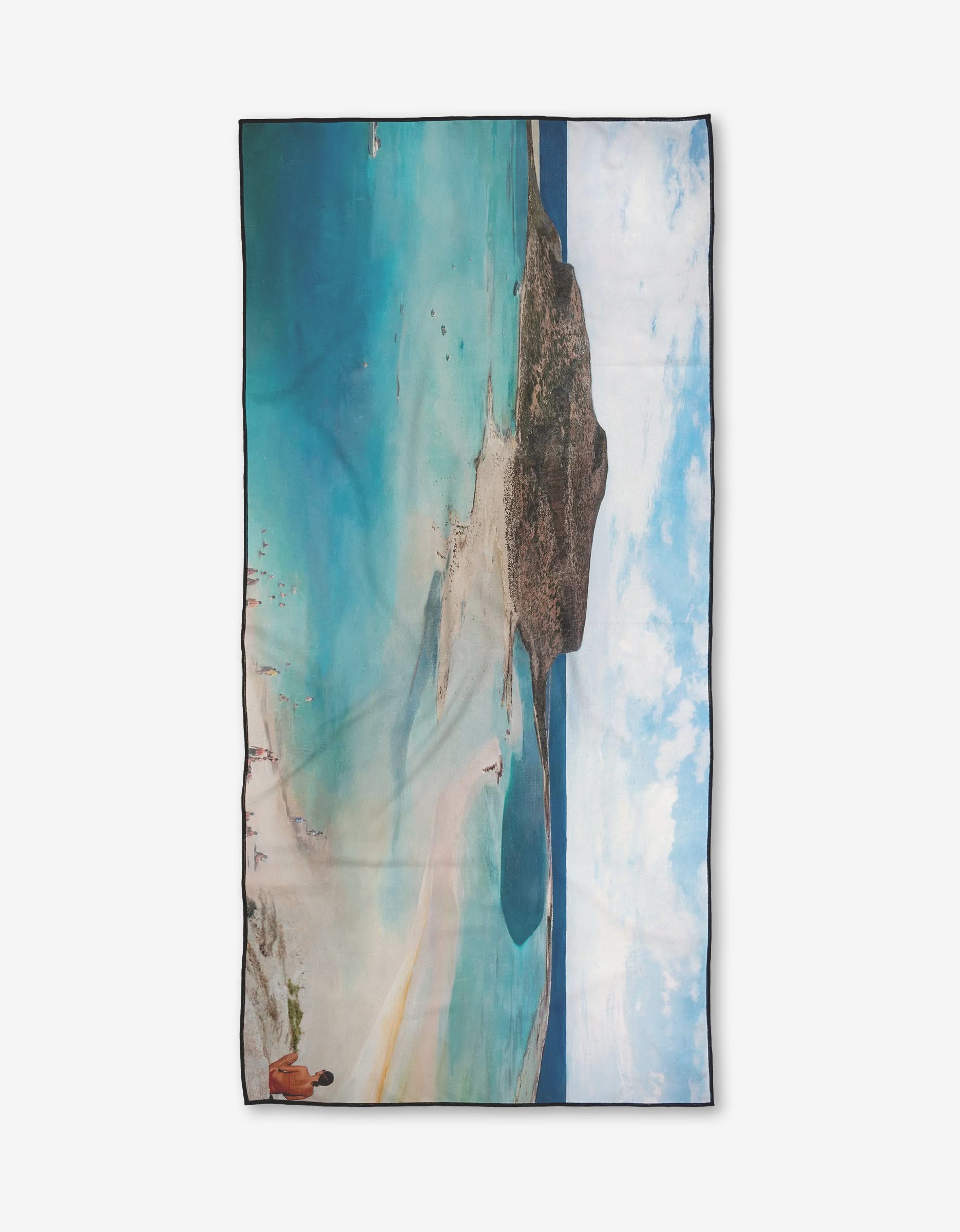 The Seaside Rendezvous beach towel by Stefanie Bales, made from recycled materials, displays an aerial image of a turquoise lagoon, sandy shore, rocky hill, and small figures under a partly cloudy sky.