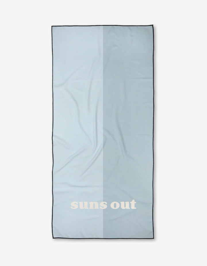 A light blue rectangular beach towel by Kelsi Fullmer, named Suns Out, features bold white suns out text near one end and is shown against a plain white background.
