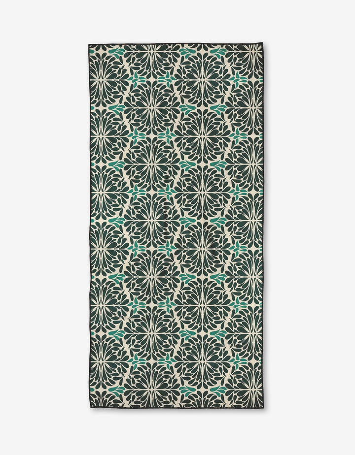 The Teal Pinwheel by Rebecca Woolbright is a rectangular micro-loop beach towel with a symmetrical floral pattern in dark green, teal, and cream, covering the entire fabric and bordered by a slim dark edge.