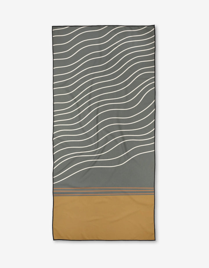 The Wavey Beach towel by Coit Creative features wavy white lines on gray with a solid mustard yellow bottom, separated by thin gray stripes. Made from recycled materials and quick-drying micro-loop fabric.