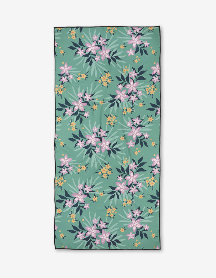 The Wilder towel by Charlie Rowan Designs is a rectangular, quick-drying towel with a green background and a pattern of pink, yellow, and white tropical flowers plus green and dark blue leaves.