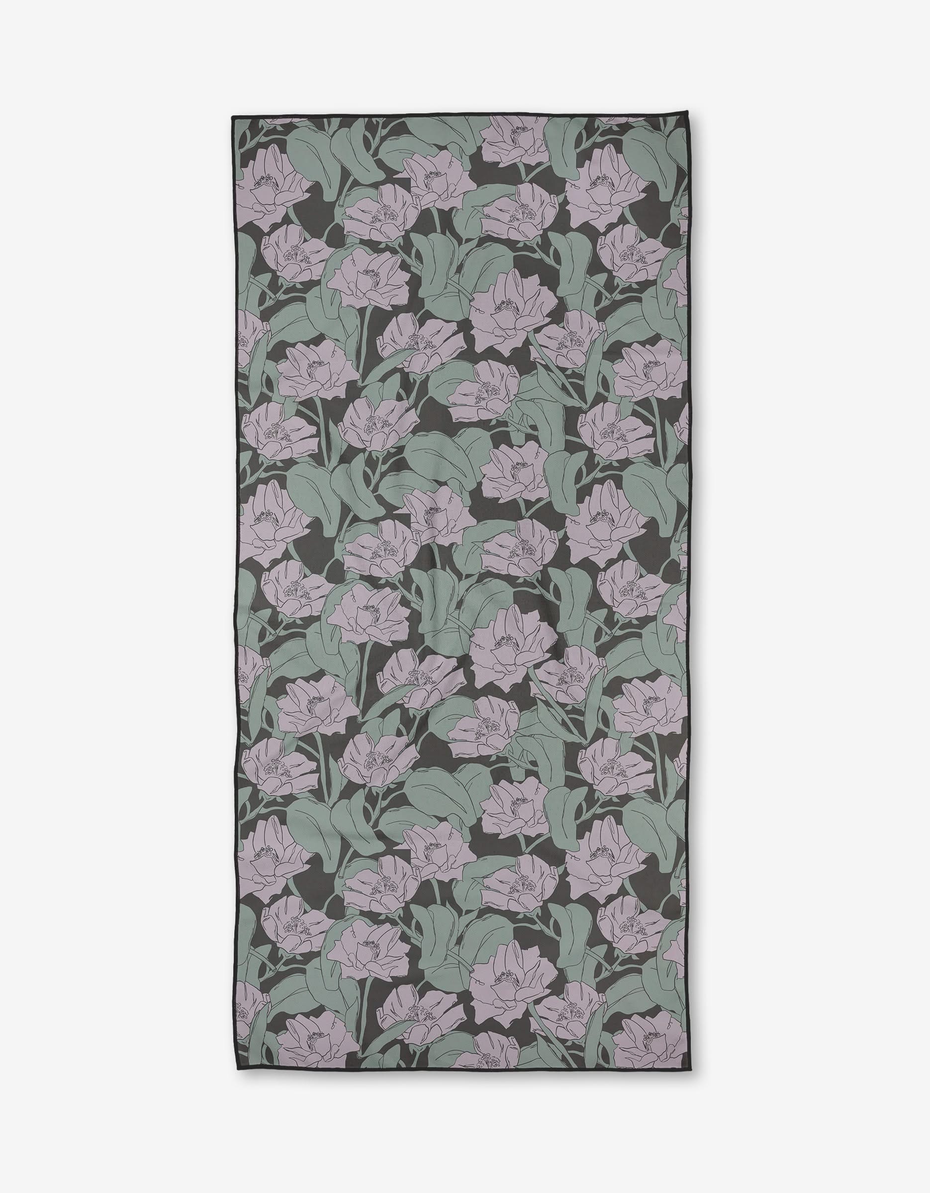 Aubrey Fairchild’s Wildflower About You is a rectangular, quick-drying towel with a floral pattern of light purple flowers and green leaves on a dark background.