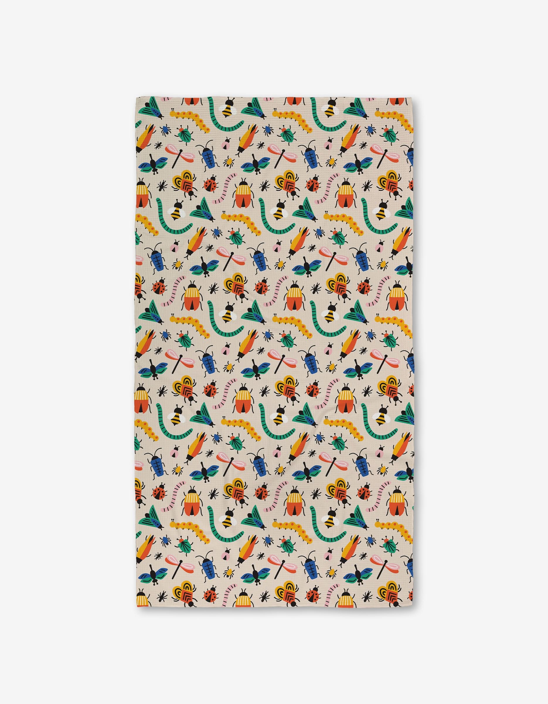 A rectangular cloth from Genna Blackburn called Bugs, made with eco-friendly towels, features a playful pattern of bugs in green, yellow, red, and blue with volcanoes, trees, and scattered leaves on a white background.