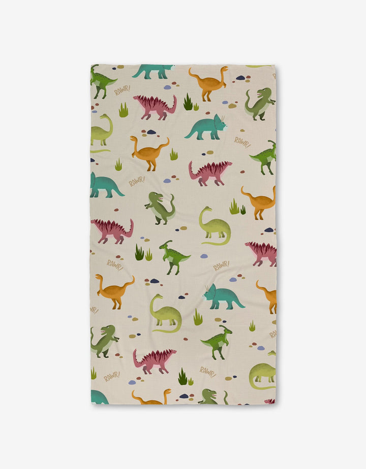 GEOMETRY’s Dancing Dinos Bath Towel, rectangular with a playful dinosaur pattern in green, orange, pink, and blue on cream with grass and rocks. Features ultra-absorbent Geoweave technology and “RAWR!” text near some dinos.