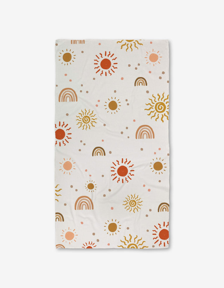 The GEOMETRY Happy Skies Luxe Bath Towel features a cheerful pattern of suns, rainbows, and dots in brown, yellow, red, and pink on ultra-absorbent fabric evenly spread across the white surface.