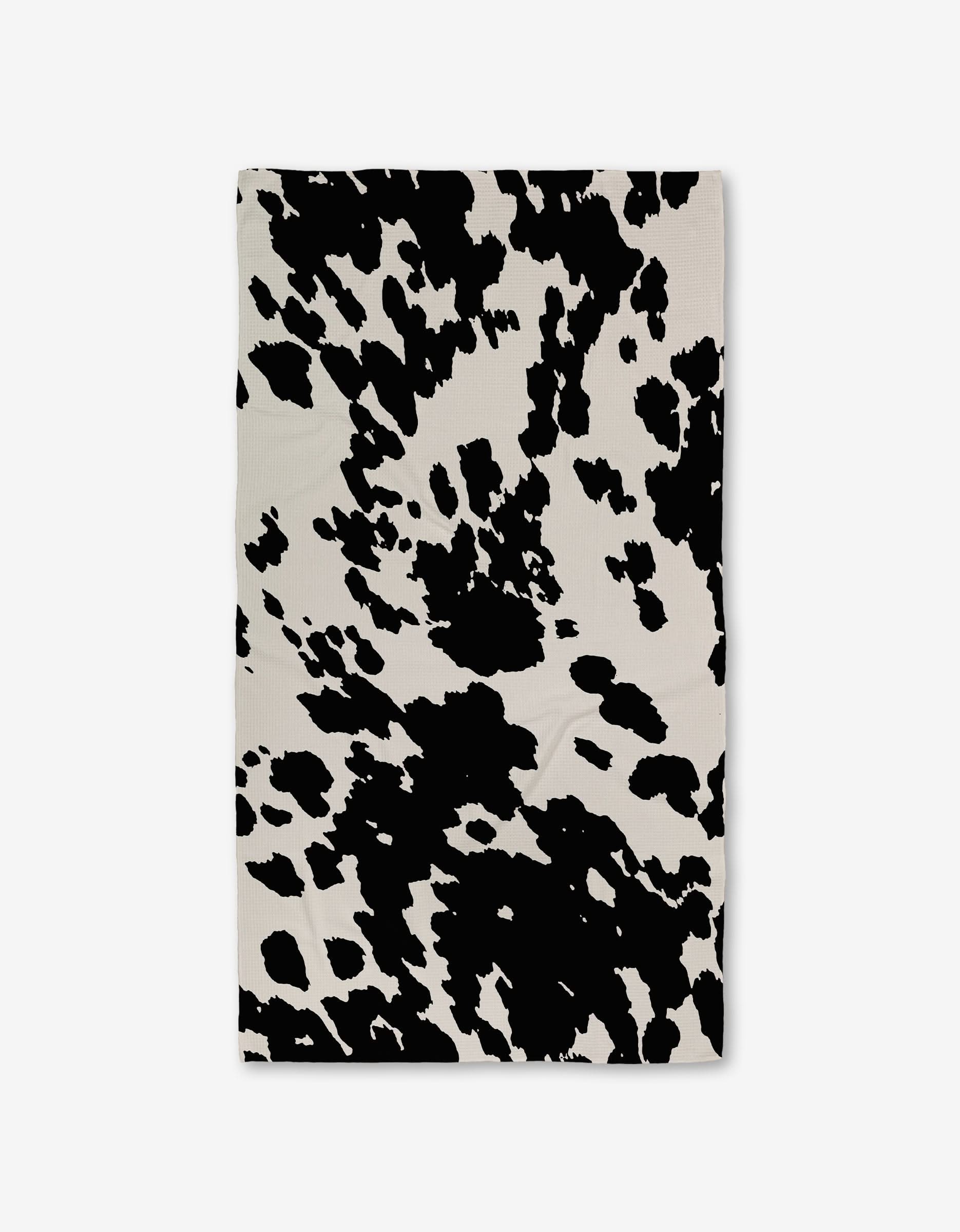 The GEOMETRY Wild West Luxe Bath Towel, featuring ultra-absorbent Geoweave technology, showcases irregular black spots on a white background in a cow print pattern and is laid flat against a plain surface.