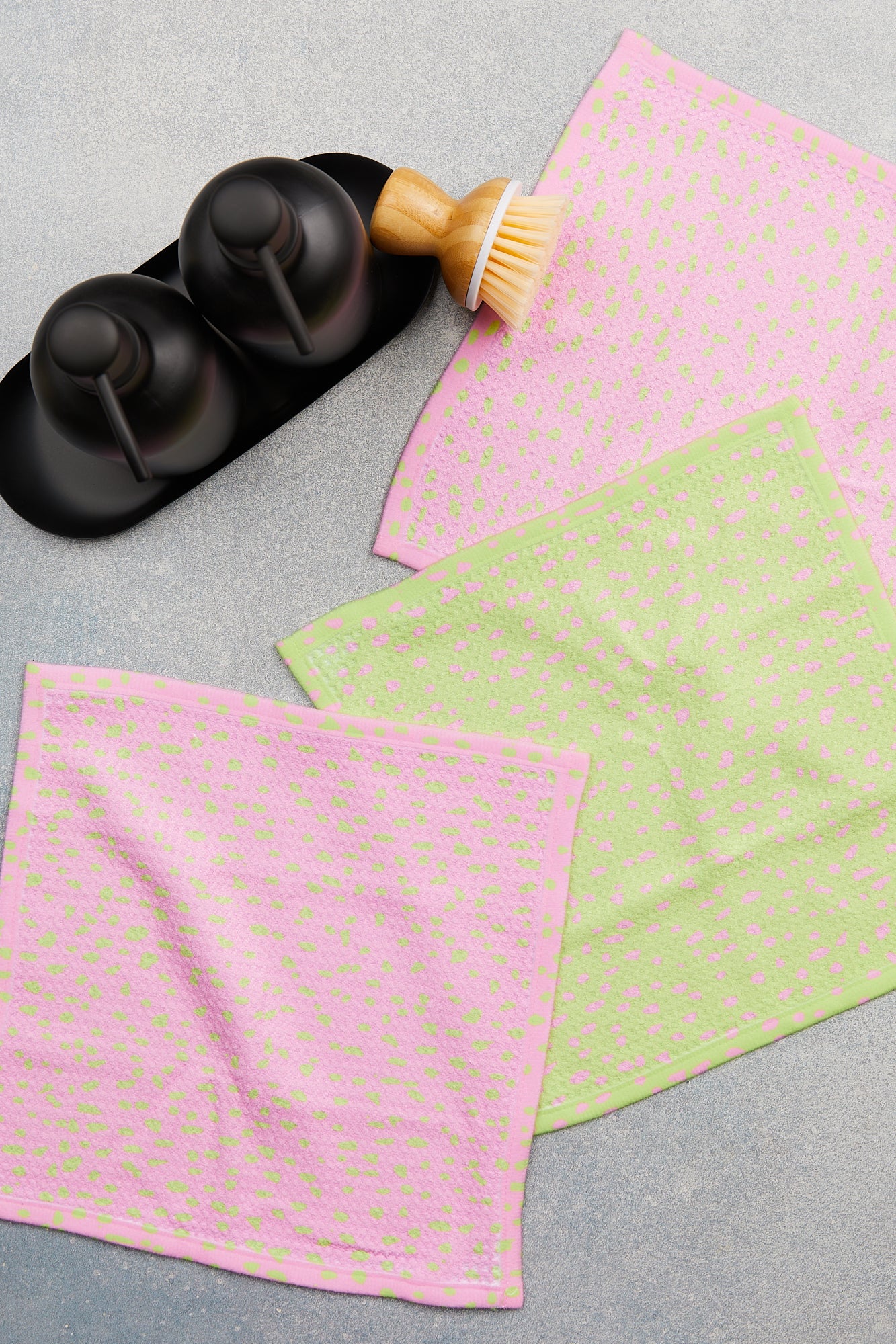 Two GEOMETRY Speckle Green Pink dishcloths (two pink, one green, all with dotted patterns and super-absorbent Geoweave fabric) lie on a light gray surface near two soap dispensers, a scrubbing brush, and a black tray.