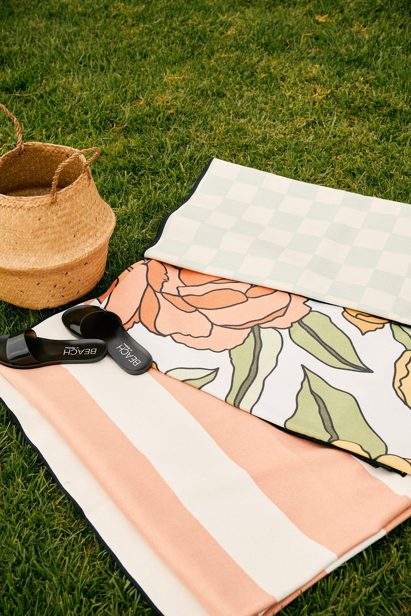 The GEOMETRY Coral Block Stripes beach towel, folded beside two other patterned towels, rests on grass next to a woven basket, a quick-drying beach blanket, and black BEACHi paddleball paddles.