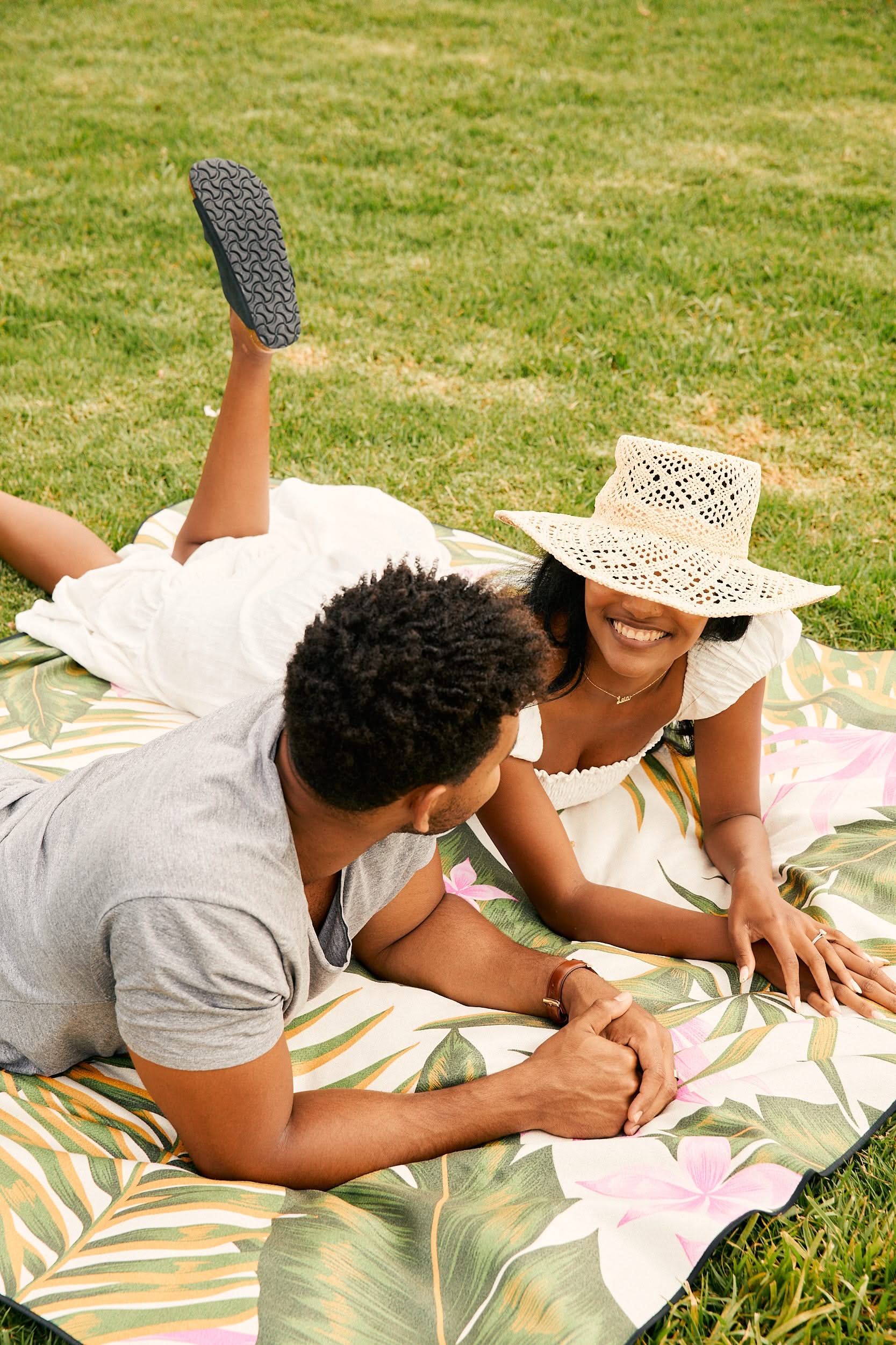 A smiling couple relaxes on the GEOMETRY Classic Checker blanket made from recycled materials, lying on green grass. The woman in a white dress and straw hat and the man in a gray shirt enjoy the outdoors together, looking content.