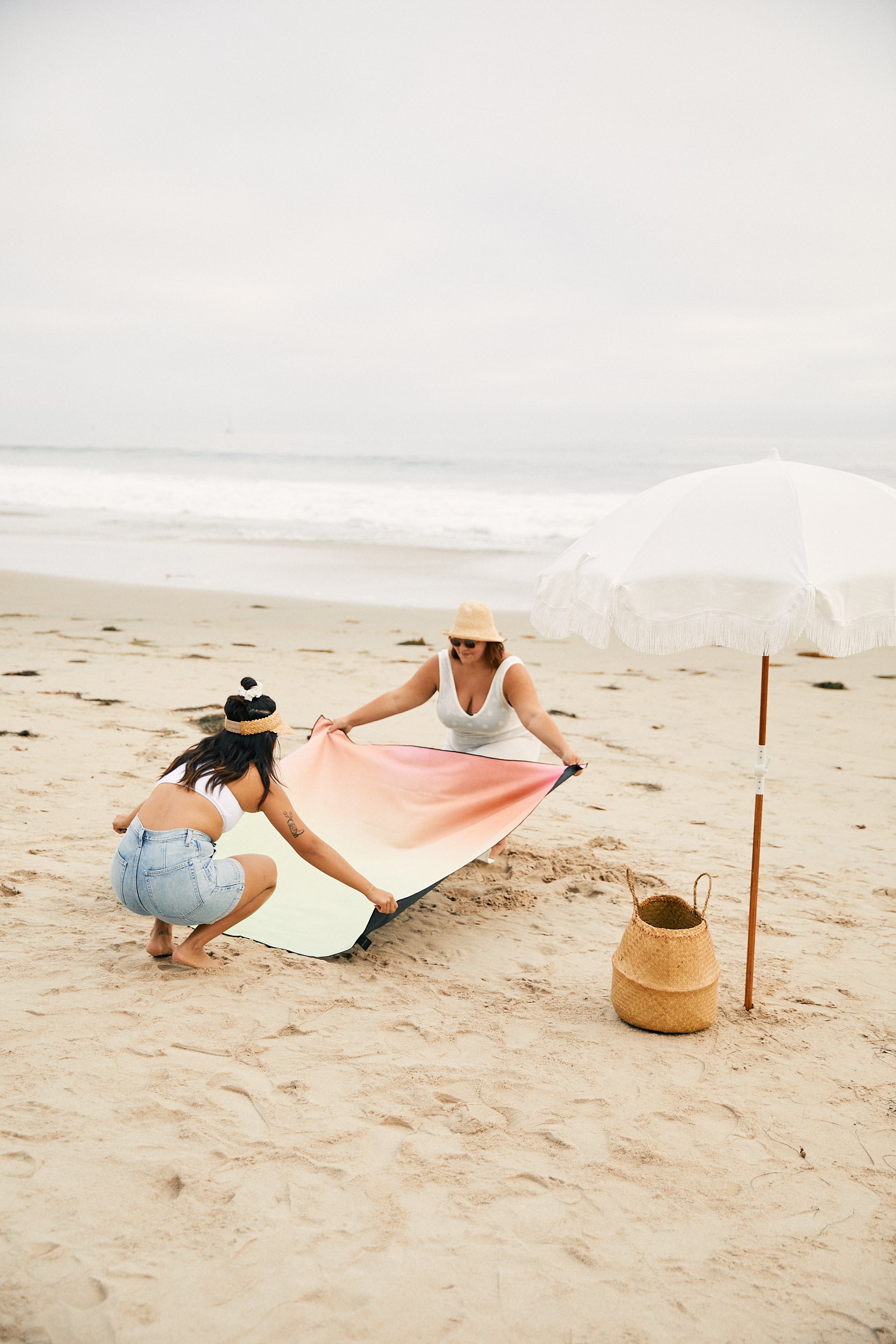 Two people are spreading the GEOMETRY Light Blue Stripes quick-drying beach blanket, made from recycled materials, on a sandy shore near the ocean. A white umbrella and woven bag sit nearby under cloudy skies.