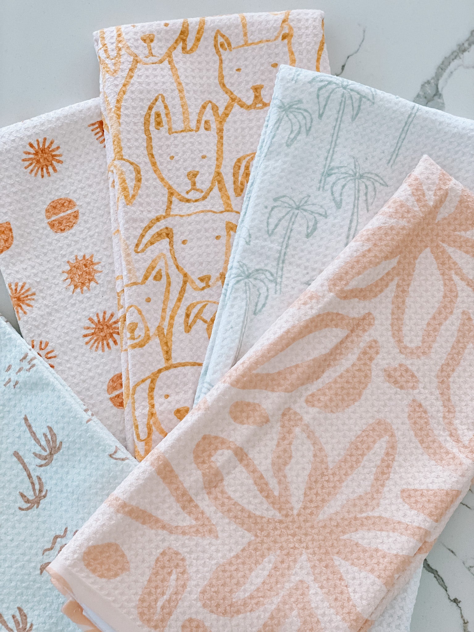 Five patterned Beach tea towels by Jen B. Peters, made from recycled materials, are fanned out on a light surface. Designs feature orange dogs, sun shapes, palm trees, small plants, and large orange flowers.