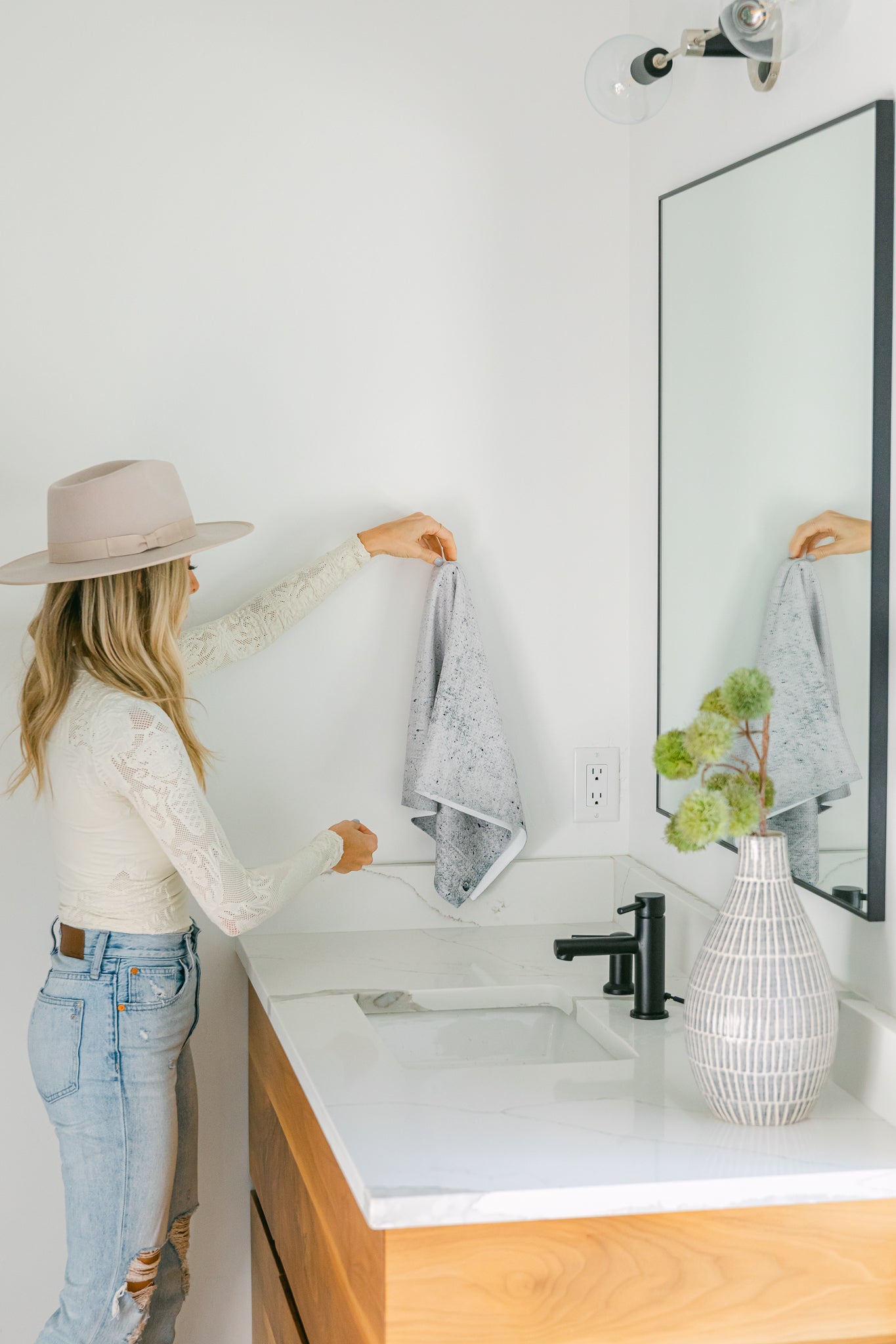 A woman in a hat and light clothes hangs the ultra-absorbent Hudson Grey towel by Kailee Wright on a hook near the bathroom sink, with a modern light wood vanity, large mirror, and white vase with greenery in view.