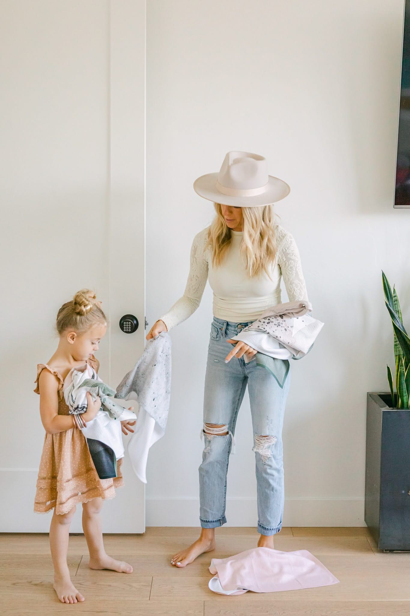 A woman in a hat and ripped jeans stands barefoot indoors with a young girl in a peach dress, both sorting clothes near a potted plant and white wall, holding the ultra-absorbent Hayden Abstract tea towel by Kailee Wright.