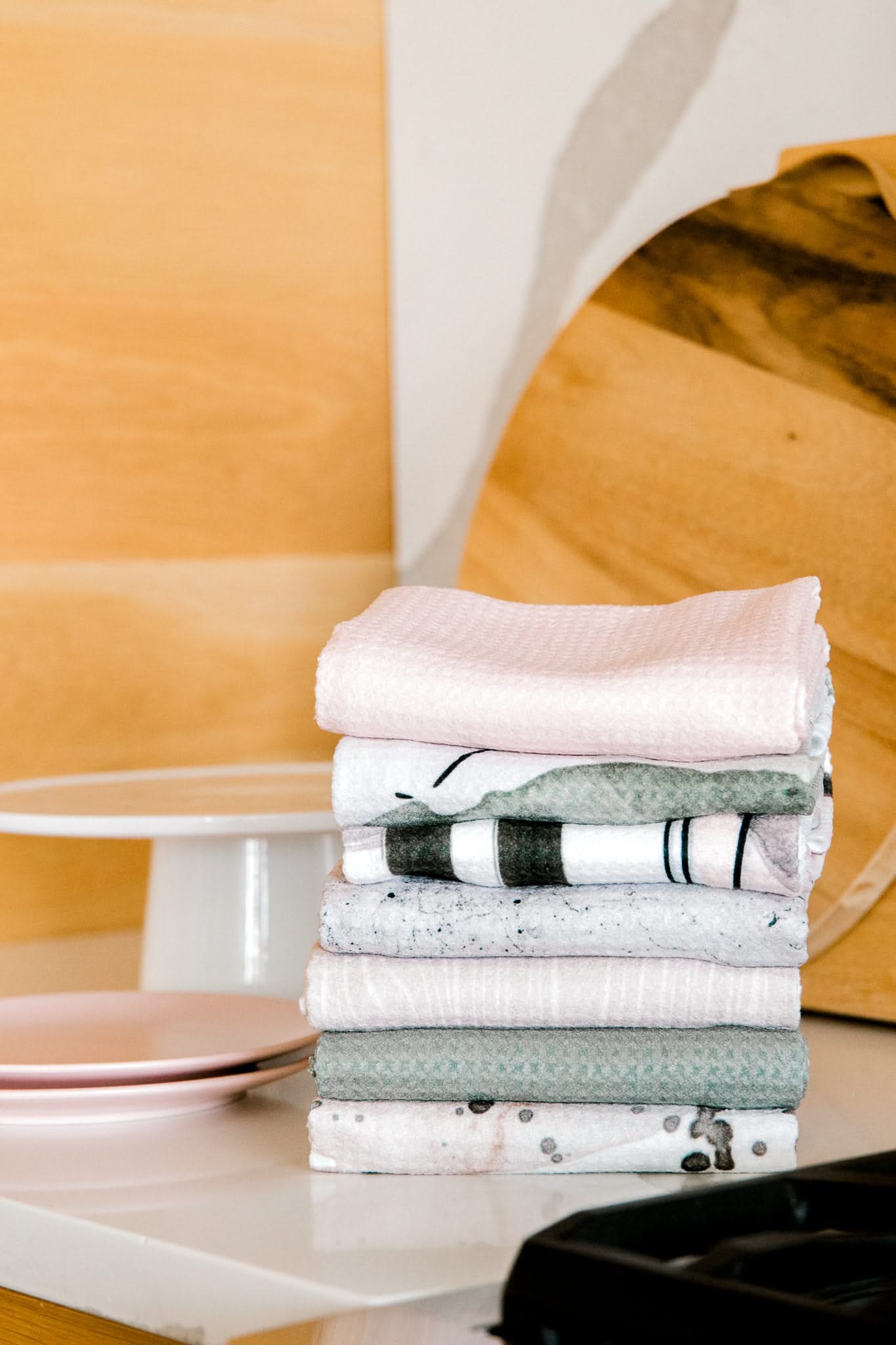 A neatly folded stack of Harper Abstract tea towels by Kailee Wright in pastel colors rests on a white countertop, with pink plates and a wooden cutting board in the background.