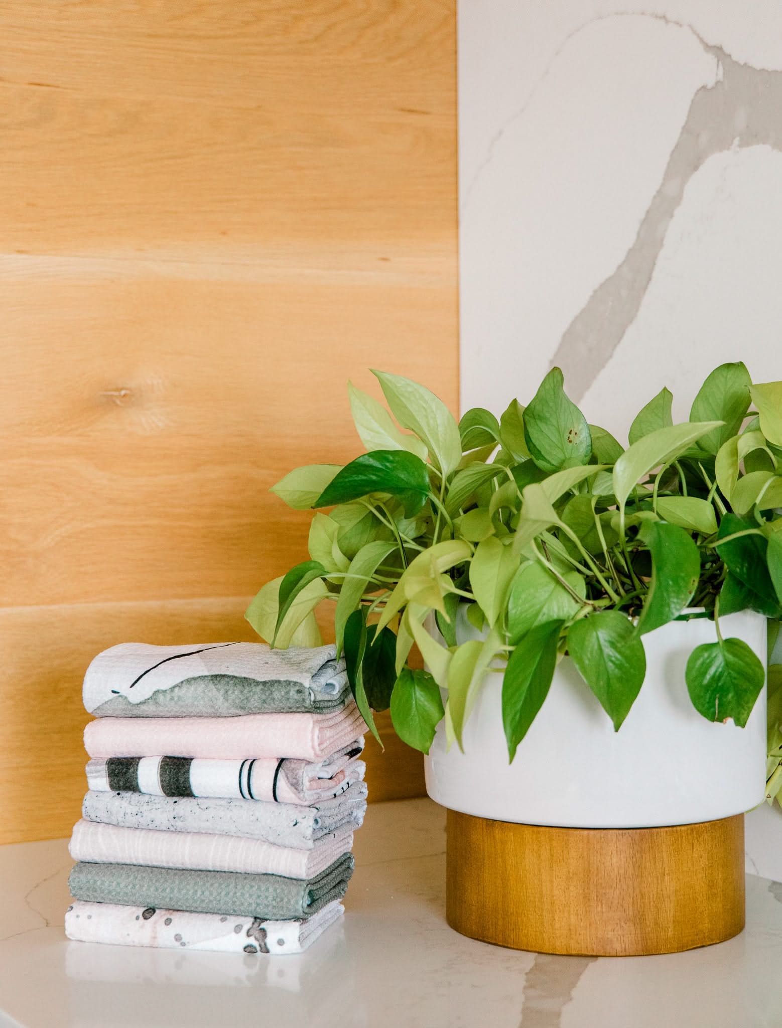 A Harper Abstract green leafy plant in a white pot sits on a wooden stand next to neatly folded Kailee Wright Geoweave Bar Towels, all arranged on a light surface with wood and marble walls in the background.