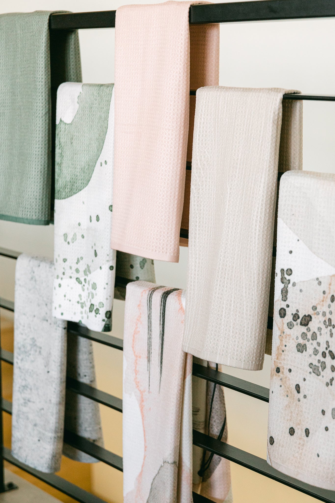 Several pastel-colored and patterned Houston Wood tea towels by Kailee Wright hang neatly over a black metal rail, showcasing ultra-absorbent Geoweave fabric in solid pink, green, beige, and abstract watercolor designs in soft hues.