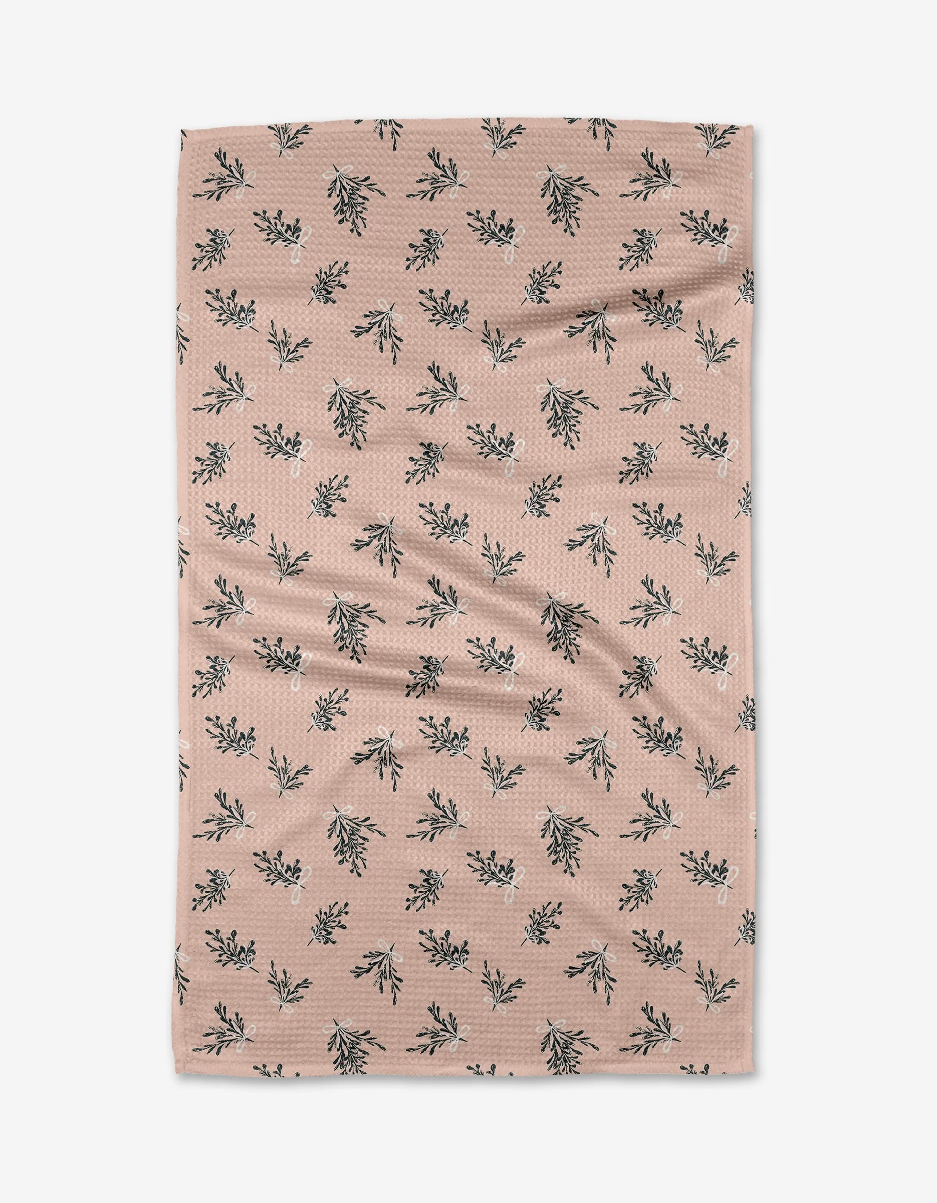The Boughs of Holly tea towel by Charlie Rowan Designs is a light brown, rectangular cloth featuring a repeating pattern of small black and white leafy branches, shown flat with slight wrinkles.