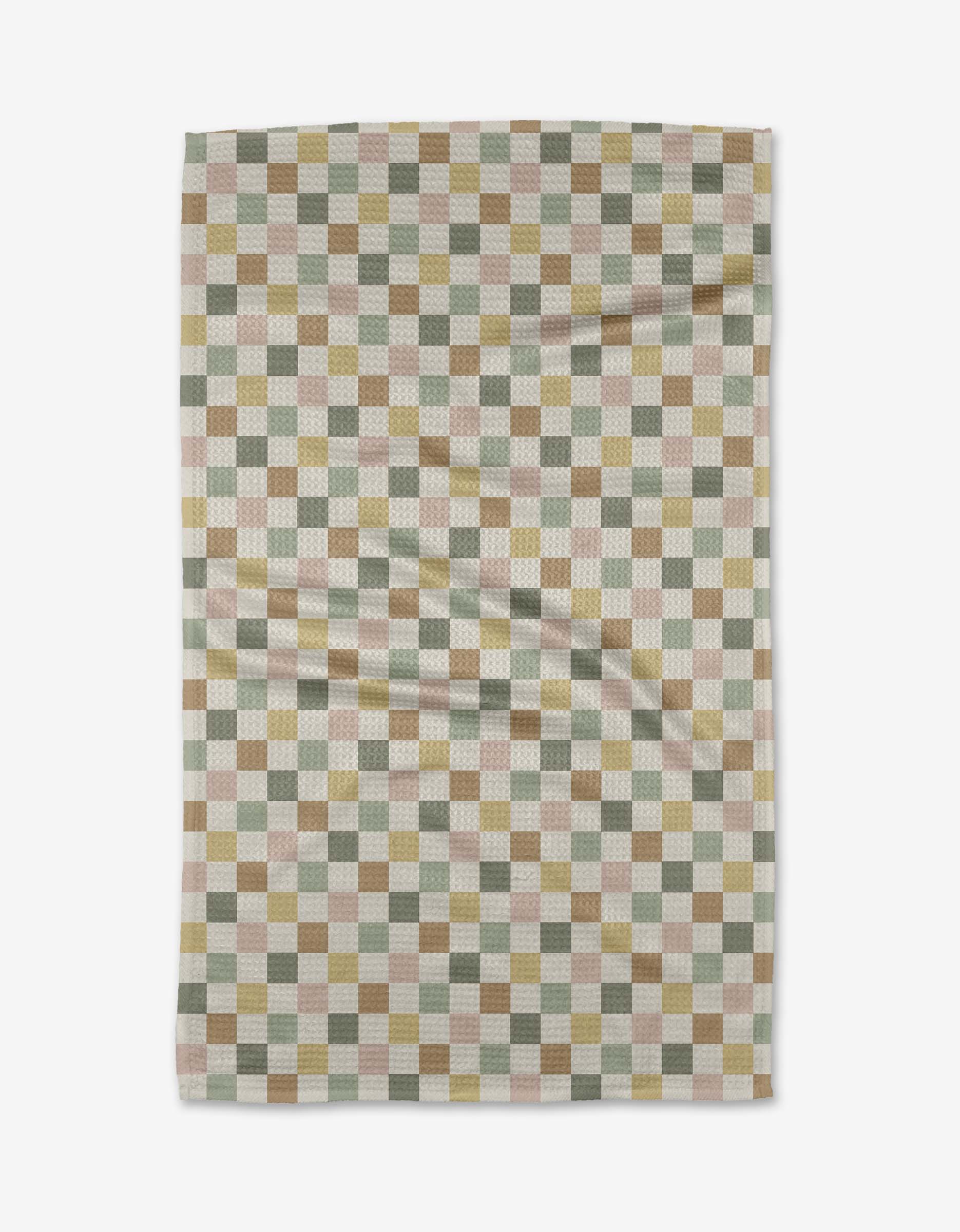 The Check Me Out tea towel by Kelsi Fullmer showcases a checkered design in muted green, tan, cream, and brown squares. Shown laid flat with visible creases.