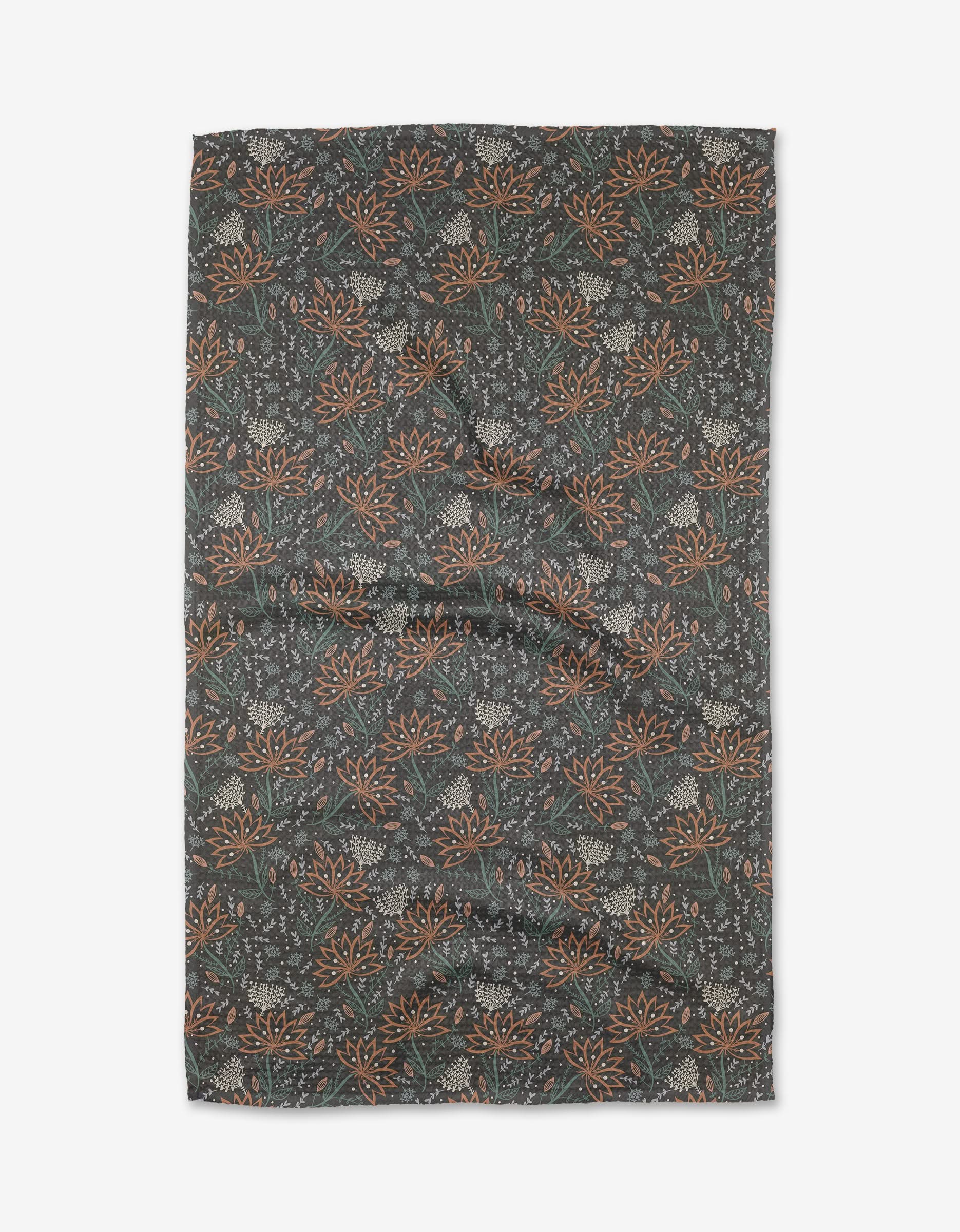 The Coral Blooms tea towel by Amita Nair features a dense orange and white floral pattern with small leaves on a dark background. Made from recycled materials, this ultra-absorbent, slightly wrinkled cloth is shown against a white backdrop.