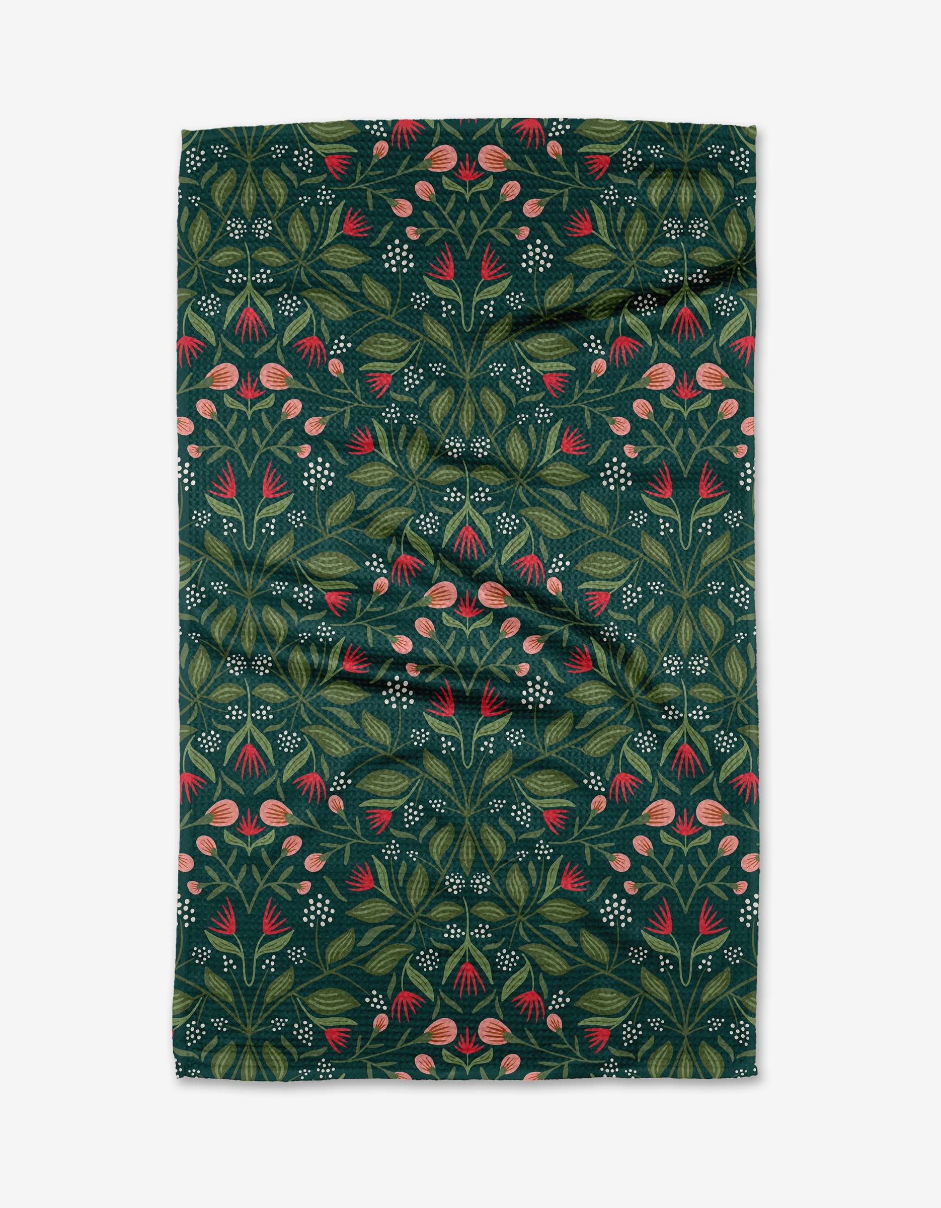 The Deck the Halls kitchen towel by Rebecca Woolbright features a dark green background with a symmetrical design of pink flowers, green leaves, and white dots. The rectangular fabric is shown laid flat with subtle wrinkles.