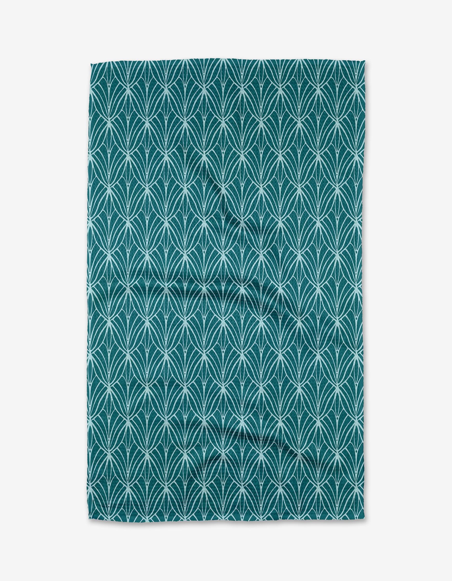 The Fish Tails kitchen towel by Marta Harvey is teal with a repeating white geometric leaf pattern and slight wrinkles visible as it’s laid flat.