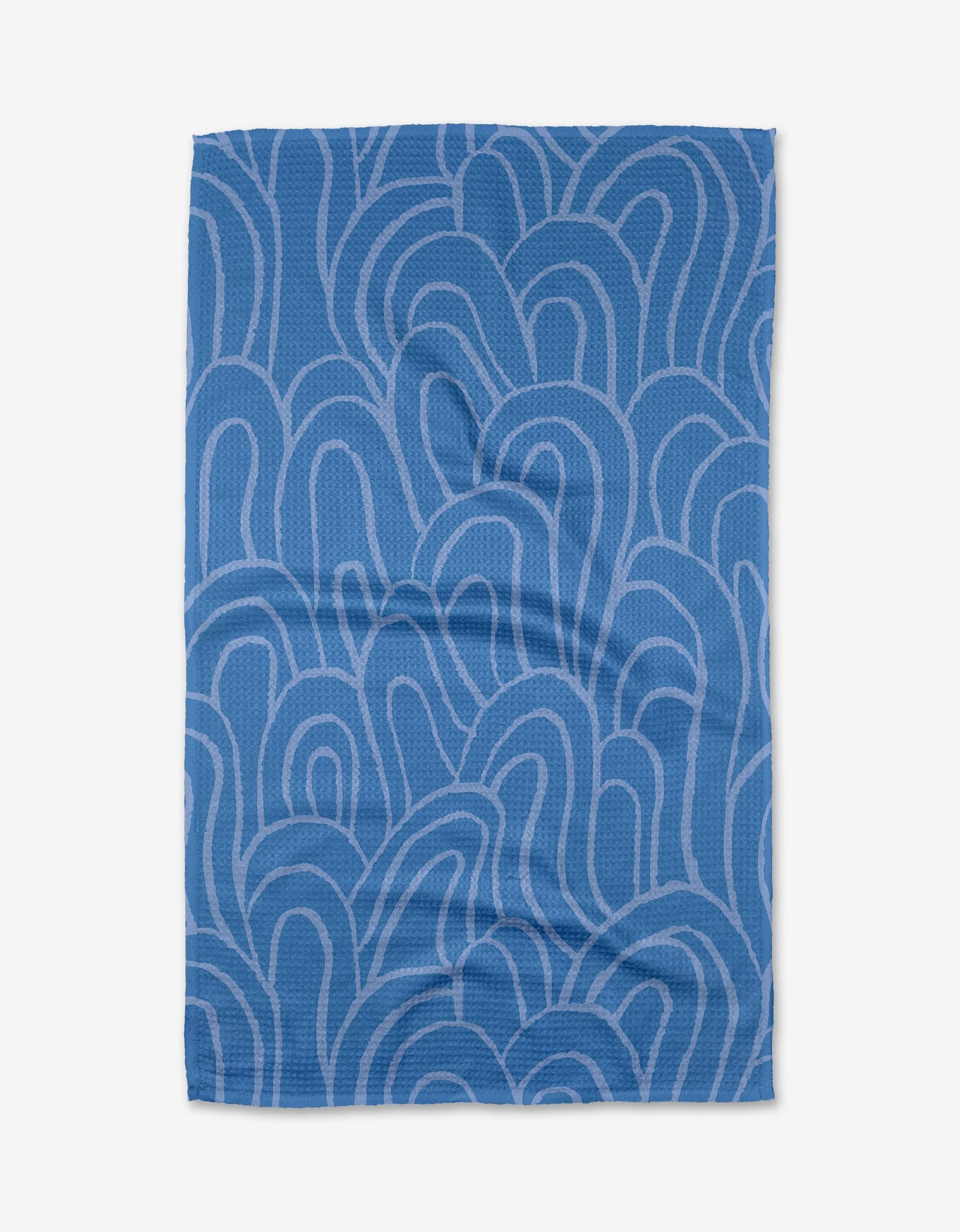 The High Tide tea towel by Sareka Unique features a blue base with wavy, abstract lighter blue lines. Made from recycled materials, it’s shown laid flat with a few visible creases.