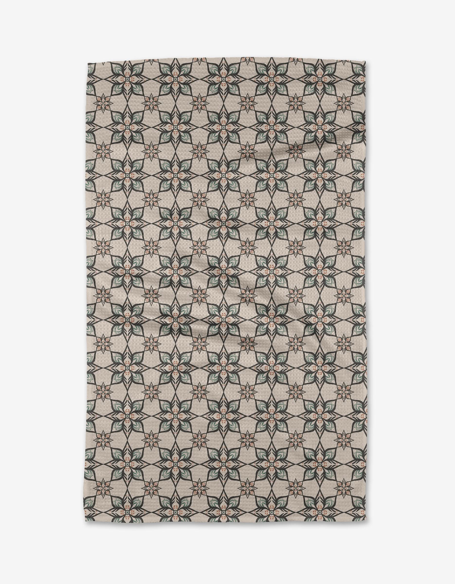 The Jungle Night tea towel by Amita Nair features a green and pink geometric floral pattern on beige Geoweave fabric, arranged in a symmetrical grid and shown slightly wrinkled against a white background.