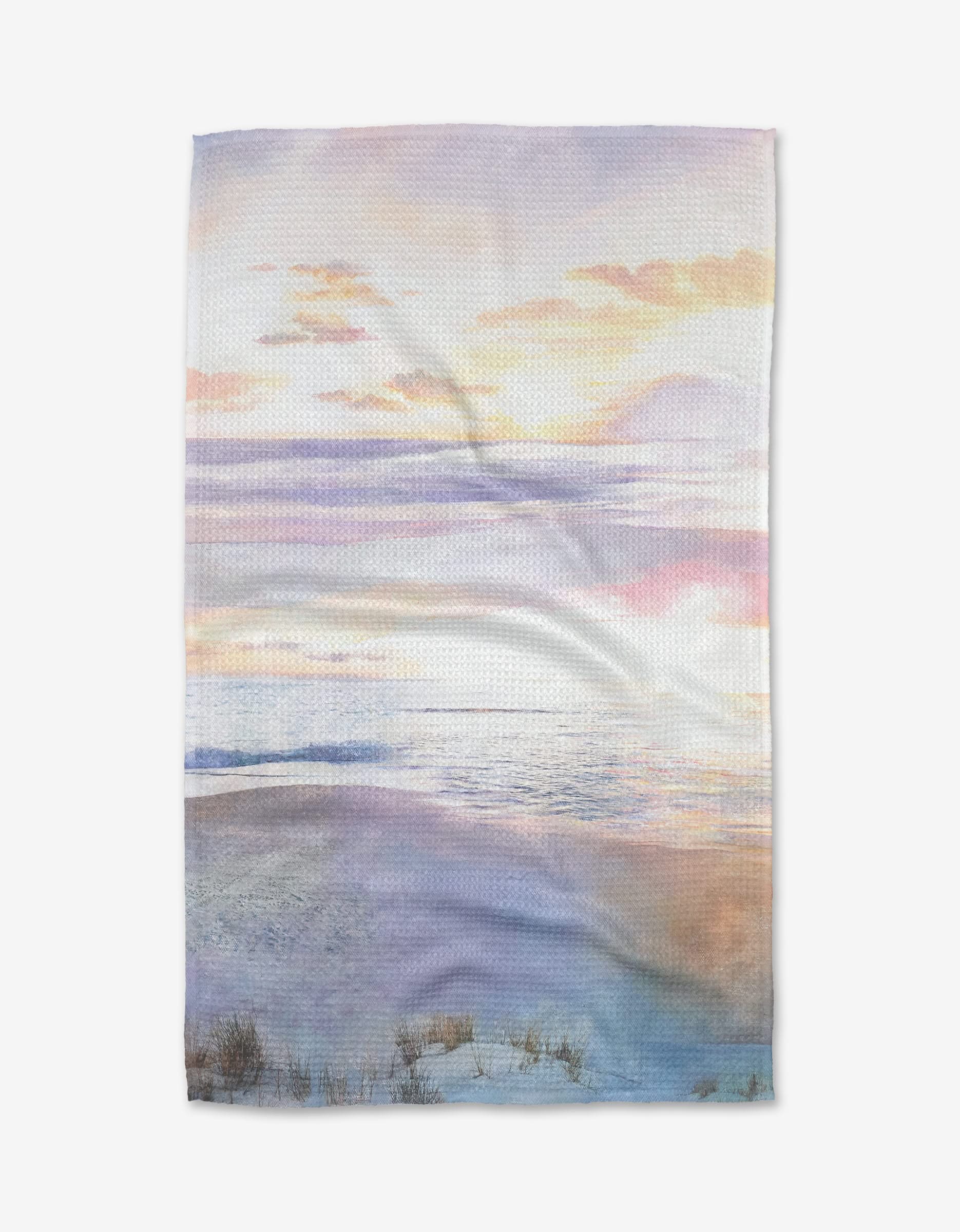 The Lilac Coast tea towel by Stefanie Bales showcases a serene beach scene with sand dunes, gentle ocean waves, and a pastel sky filled with pink, purple, and yellow sunset or sunrise clouds.
