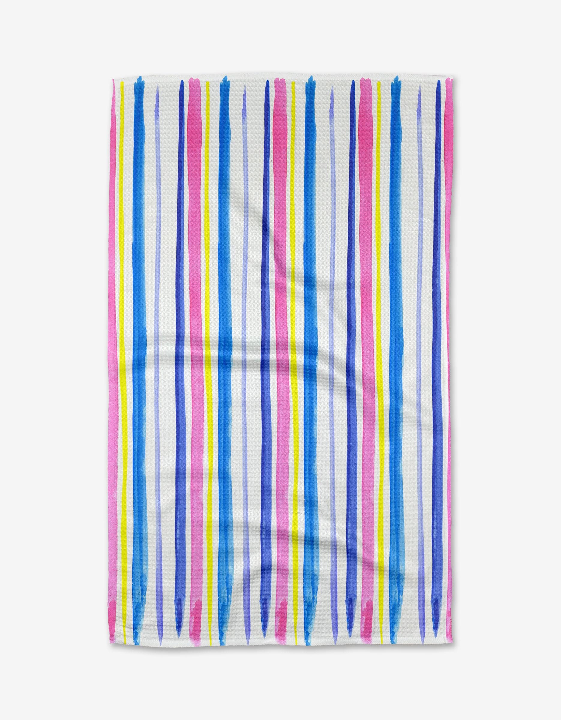 The Macarons in Paris Stripe kitchen towel by Travel Write Draw (Meagan Morrison) features brushstroke-style vertical stripes in blue, pink, purple, and yellow on a white background with a hand-painted look and visible fabric creases.