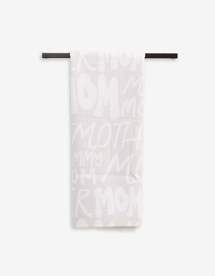 A light-colored, ultra-absorbent GEOMETRY Mom tea towel hangs on a black rod, showcasing playful white lettering with words like MOM, MOTHER, and MOMMY.