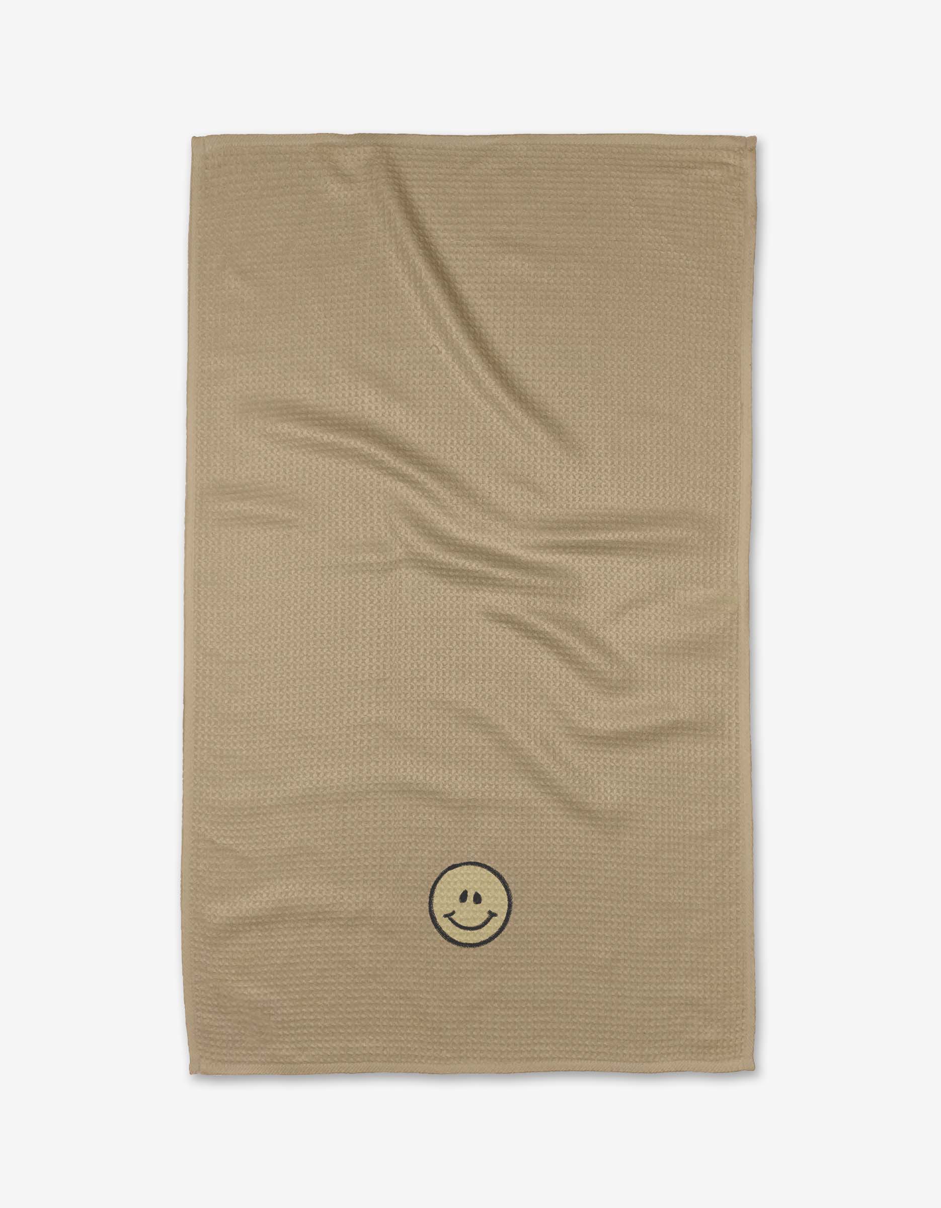 The My Happy Place tea towel by Kelsi Fullmer is beige, rectangular, made from absorbent recycled materials, and features a subtle texture and a black smiley face in one corner, shown on a white background.