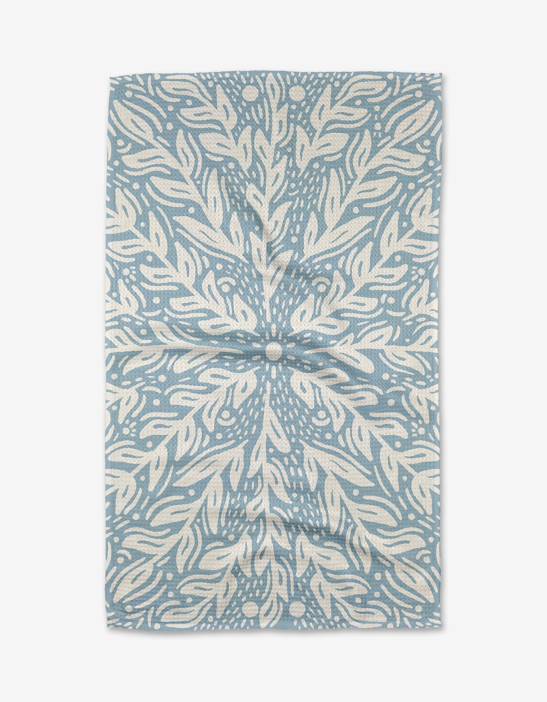 The Outwards tea towel by Graphics and Grain is rectangular, blue and white, and showcases a symmetrical botanical-inspired pattern with swirling leaves and dotted accents radiating from the center.