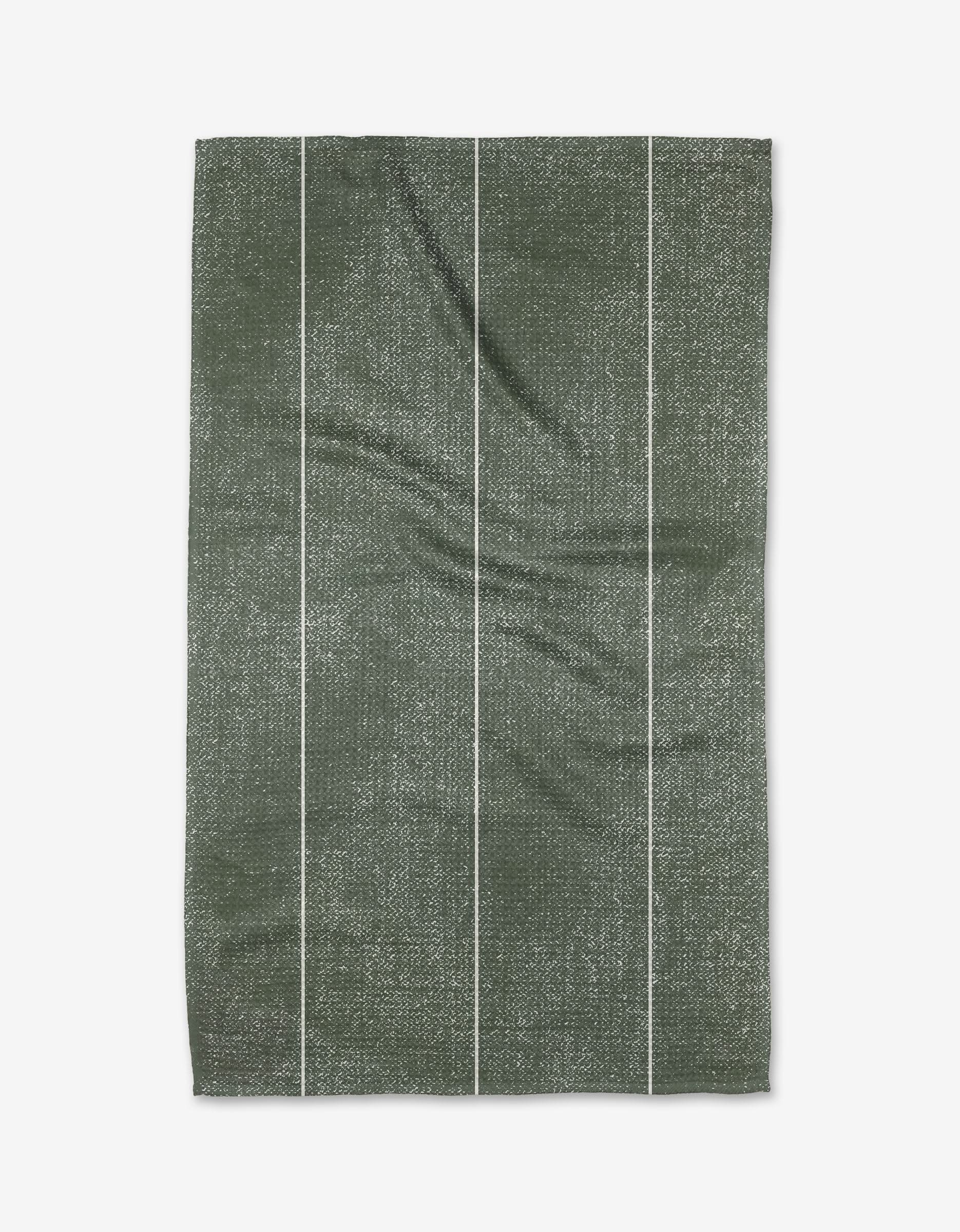 The Becki Owens Winter Pine tea towel features a rectangular shape, green color, textured pattern, and four thin white vertical stripes. Made from recycled materials, it is displayed laid flat on a white background.
