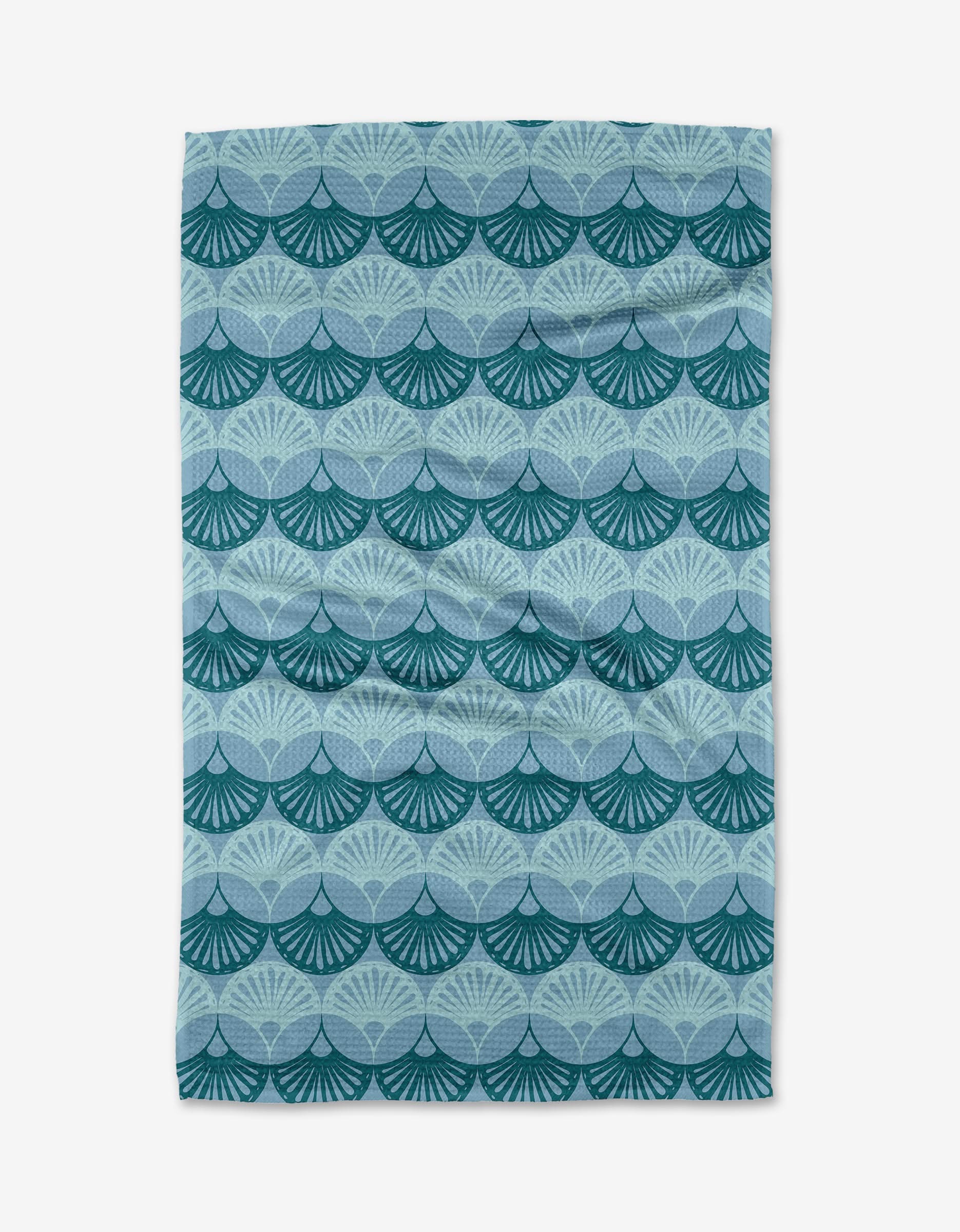 The Seashells tea towel by Marta Harvey is a rectangular Geoweave fabric made from recycled materials, featuring a repeating pattern of overlapping blue and teal fan shapes on a light blue background.
