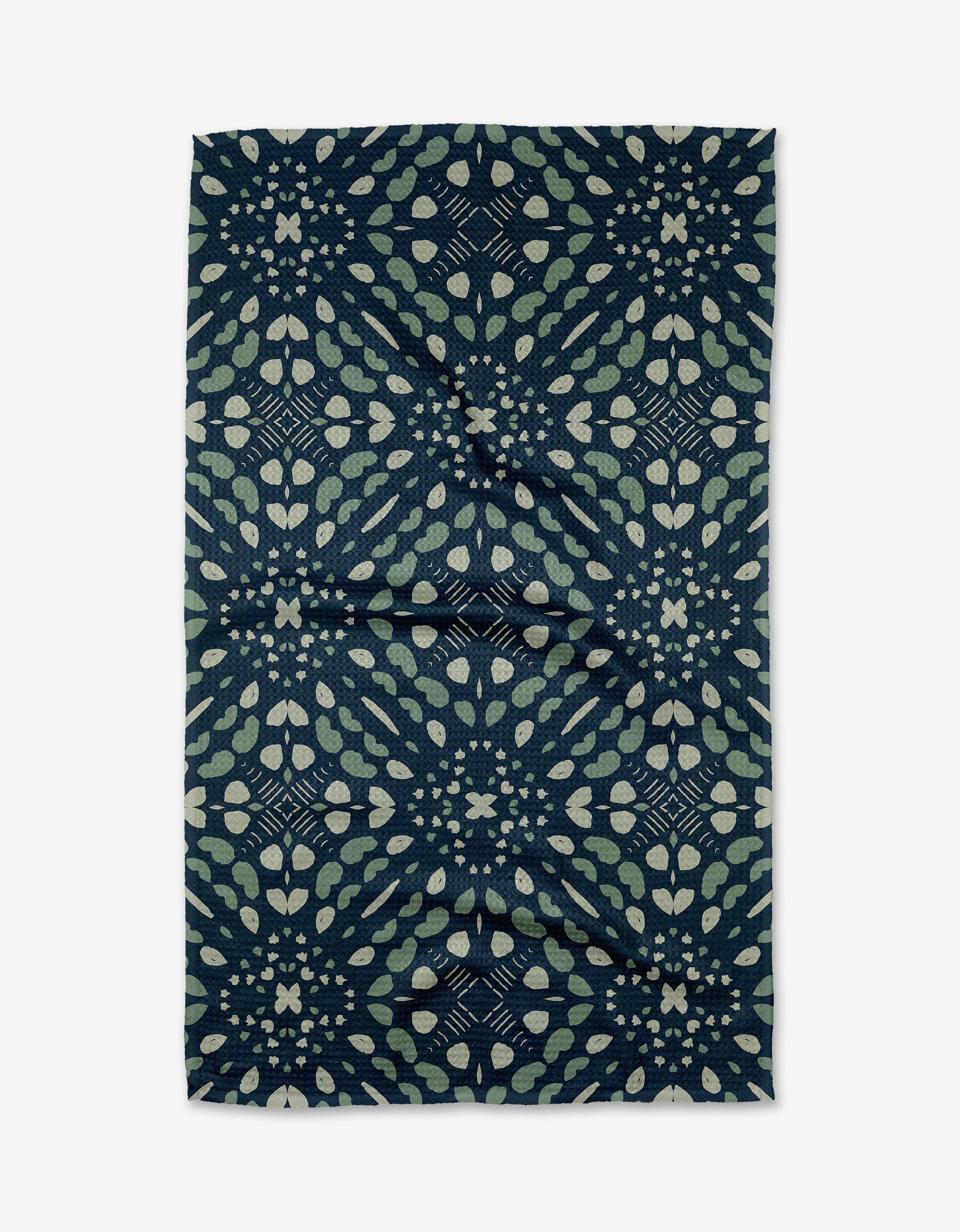 The Seeds and Nuts tea towel by Twig + Spark features a dark blue Geoweave™ fabric with light green and beige geometric leaf and petal patterns. The design is symmetrical, with the fabric showing a slightly wrinkled texture.